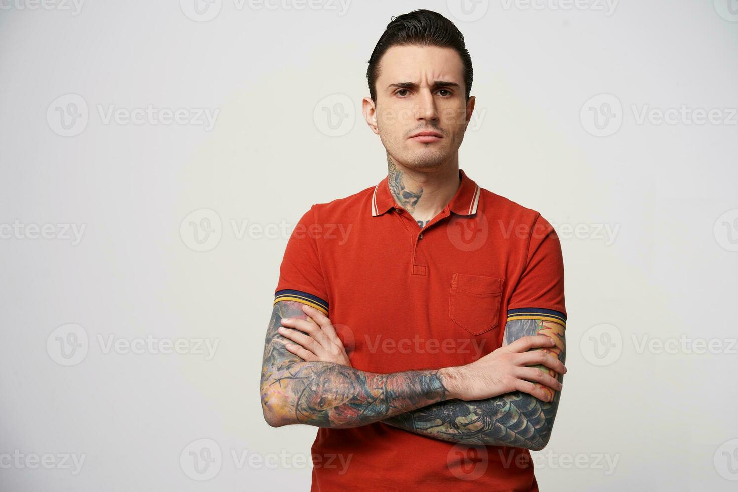 Serious man with arms folded look suspicious, eyebrows frowning, has a tattoo on his arms and neck, standing isolated on a white background and looking at camera photo