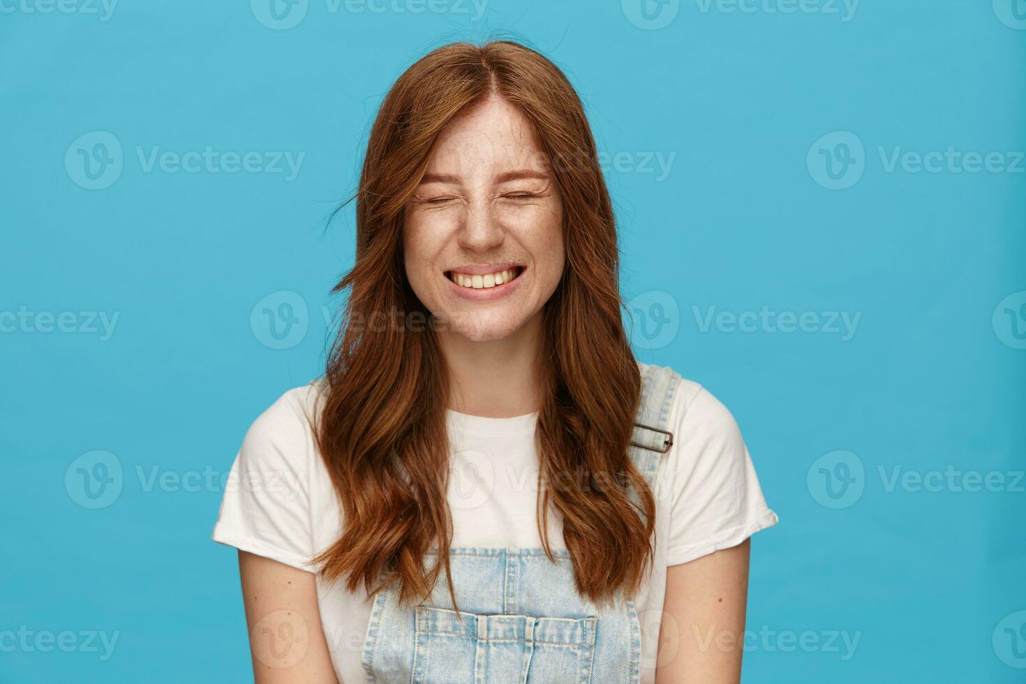 Pleasant looking young redhead happy female keeping her eyes closed while smiling happily, being in high spirit while standing over blue background in casual wear photo