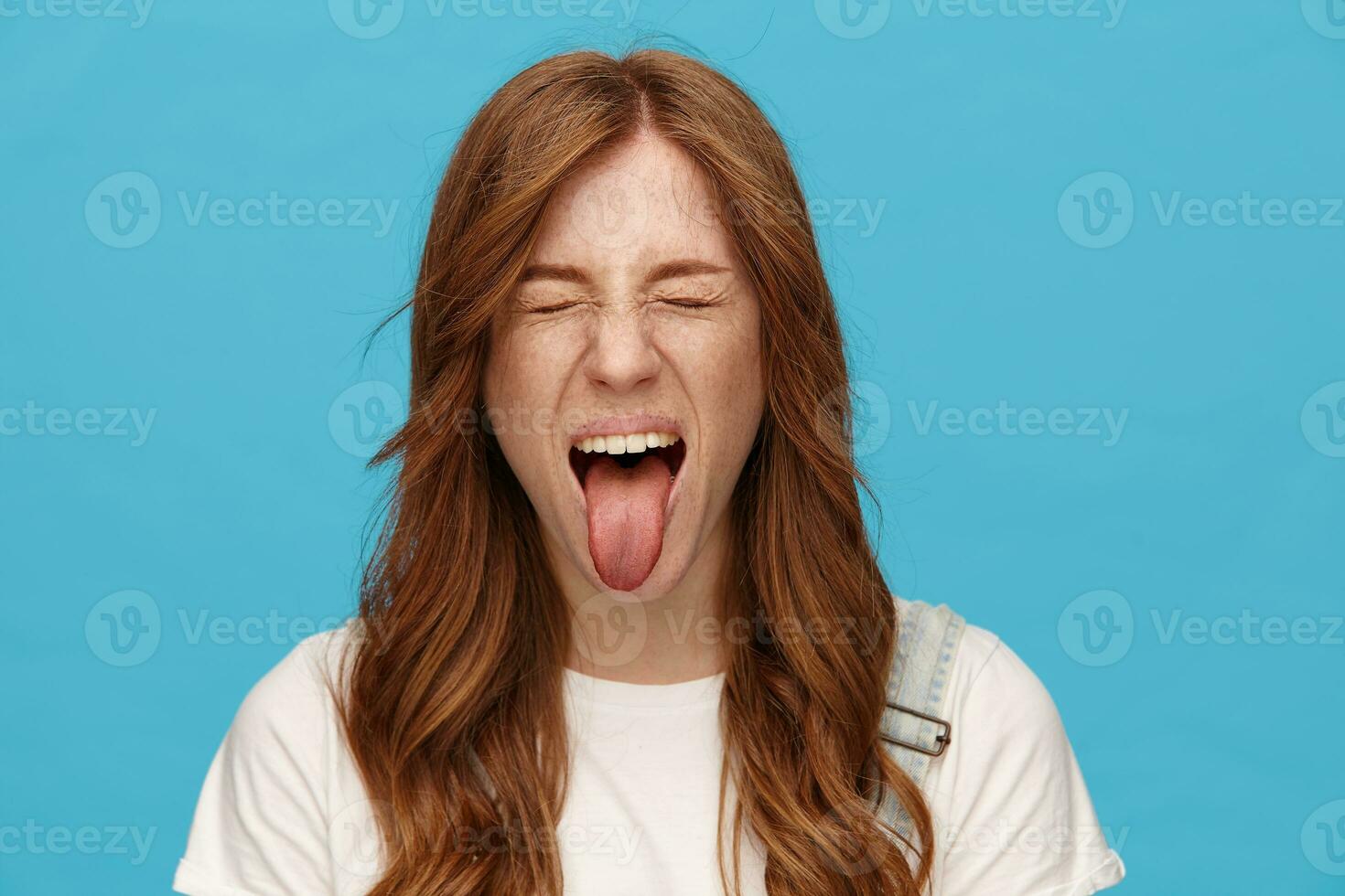 Funny shot ot young joyful redhead lady with natural makeup fooling while posing over blue background, frowning face and sticking out her tongue with closed eyes photo