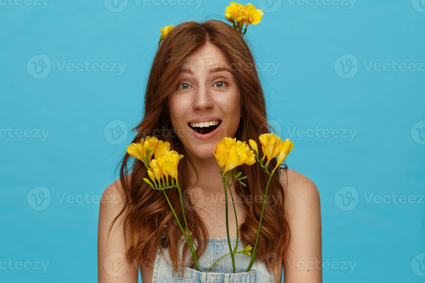 Exited young lovely lady with natural makeup raising surprisedly eyebrows while looking joyfully at camera, standing over blue background with yellow freesia photo