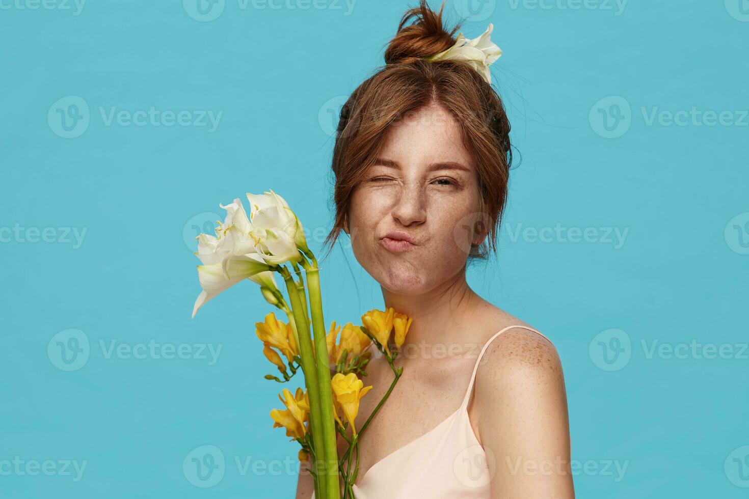 Portrait of young lovely redhead woman with natural makeup keeping one eye closed and pursing her lips while looking at camera, posing over blue background with flowers photo