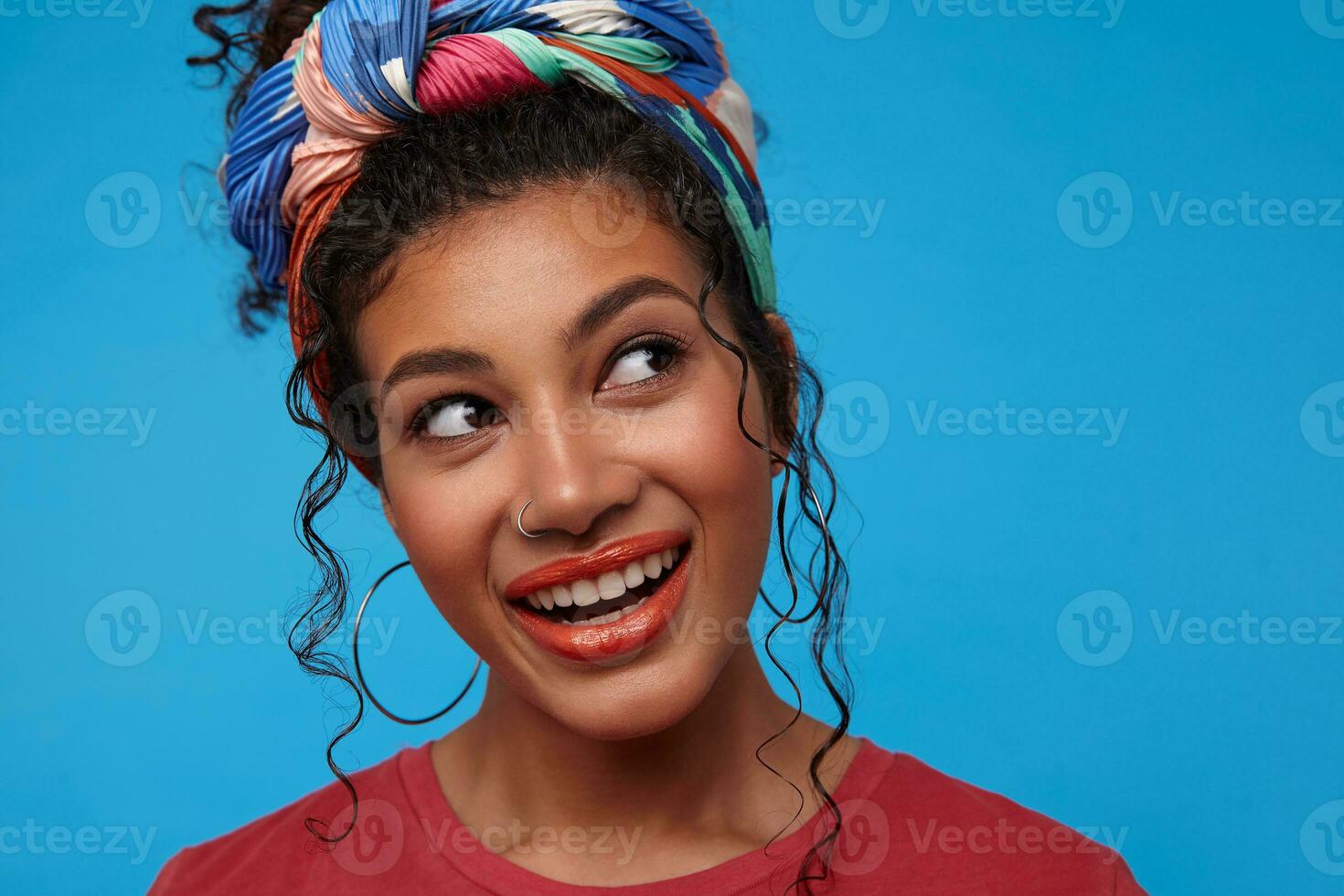 Portrait of lovely brown-eyed young dark haired curly female smiling widely while looking wonderingly aside, wearing multi-colored headband while posing over blue background photo