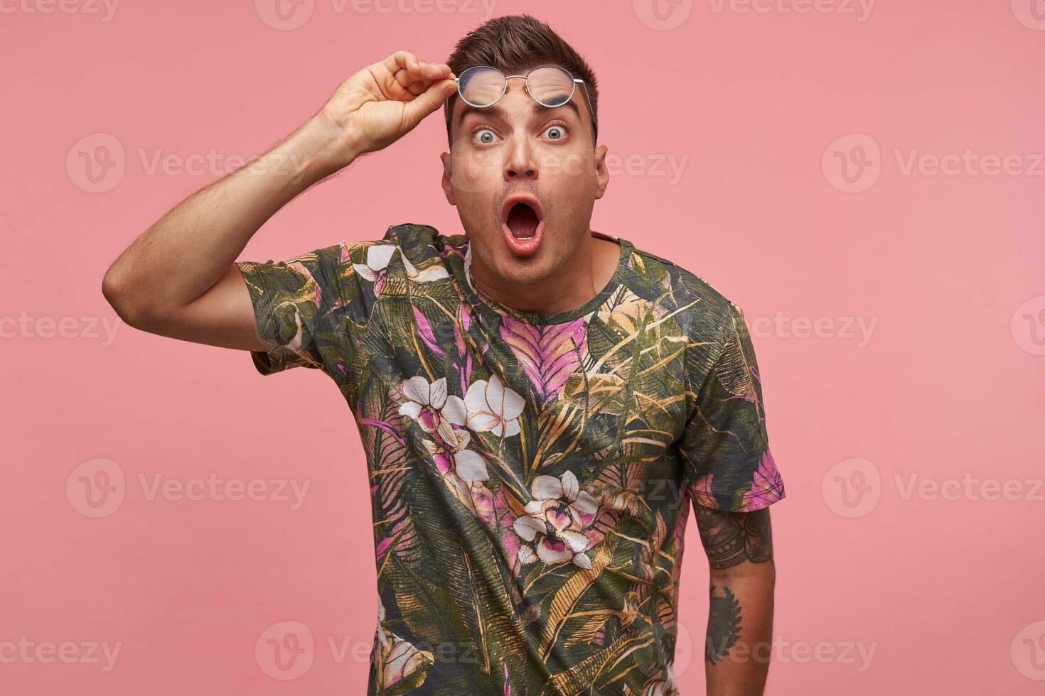 Amazed pretty young man standing over pink background with broadly eyes and mouth open, putting his glasses on forehead, wearing flowered t-shirt photo