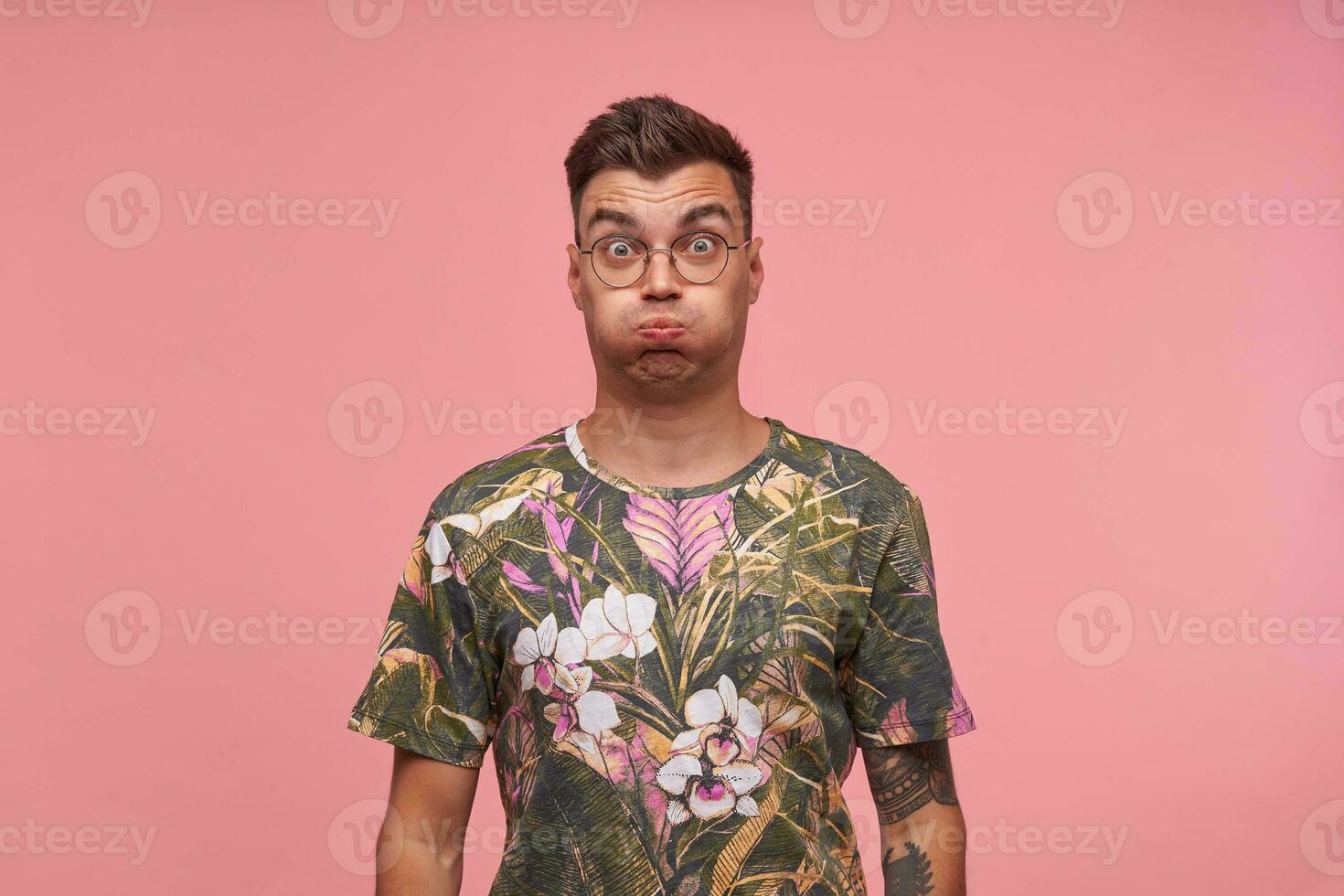 Indoor shot of cute guy in flowered t-shirt with wide eyes open standing over pink background, trying to hold breath, making funny grimace photo