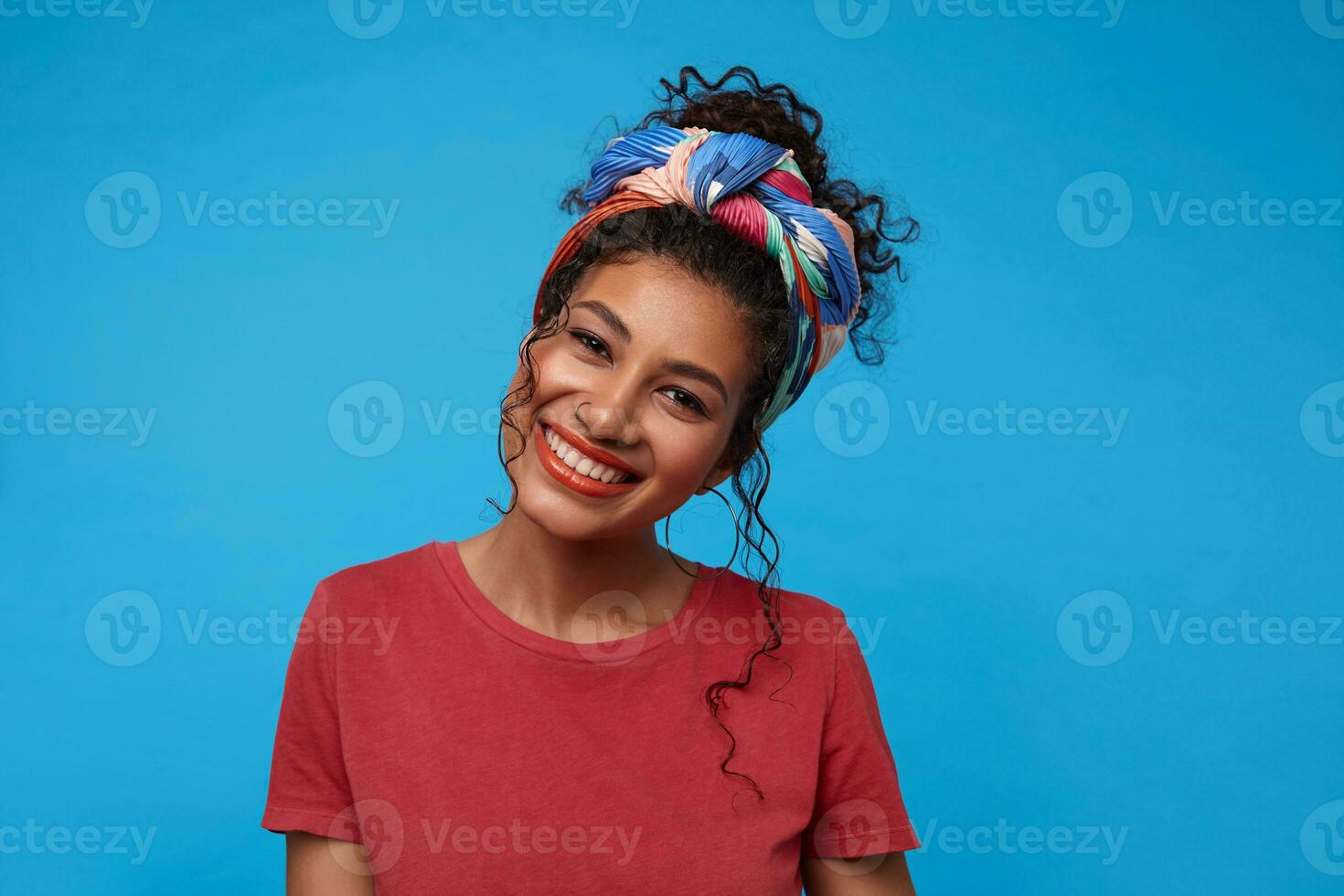 Happy young cute brunette curly female with nose piercing showing her pleasant emotions and smiling cheerfully while looking at camera, isolated over blue background photo