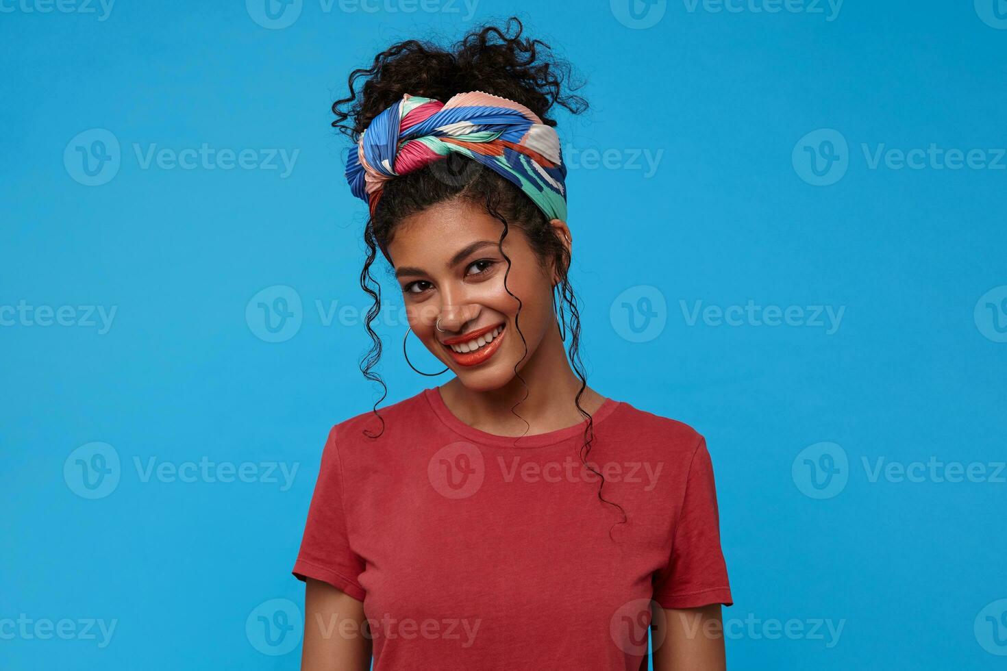 Studio photo of young lovely brunette curly woman with festive makeup being in nice mood and smiling gladly at camera, standing over blue background