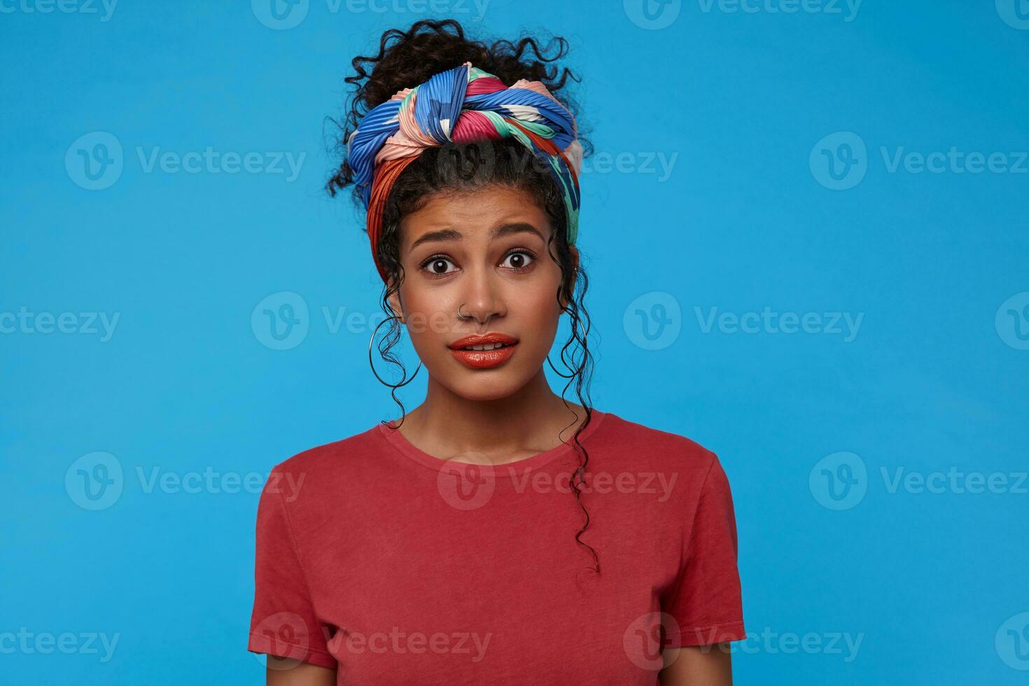Bewildered young attractive dark haired curly woman with multi-colored headband frowning eyebrows while looking confusedly at camera, isolated over blue background photo