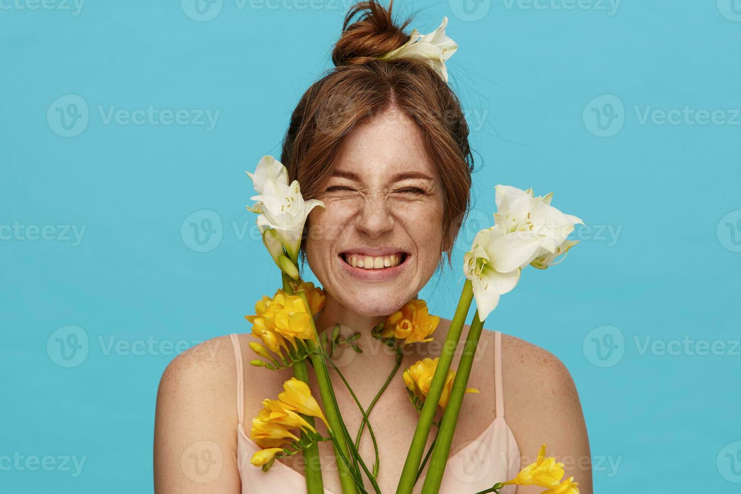Studio shot of young joyful redhead female with bun hairstyle frowning happily her face while smiling widely, isolated over blue background with bunch of flowers photo