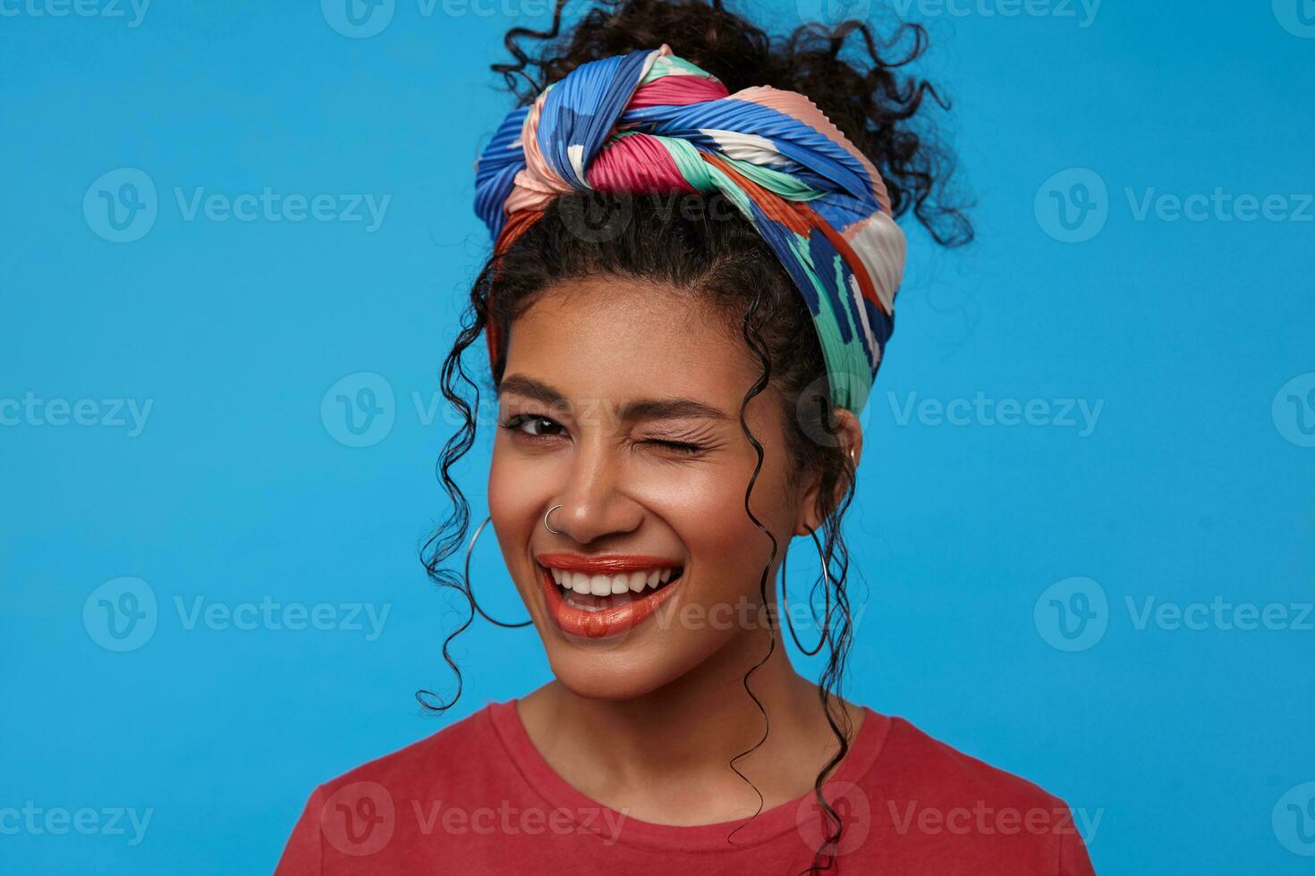 Joyful young attractive brunette curly woman with festive makeup winking cheerfully at camera while smiling gladly, standing over blue background in colored clothes photo