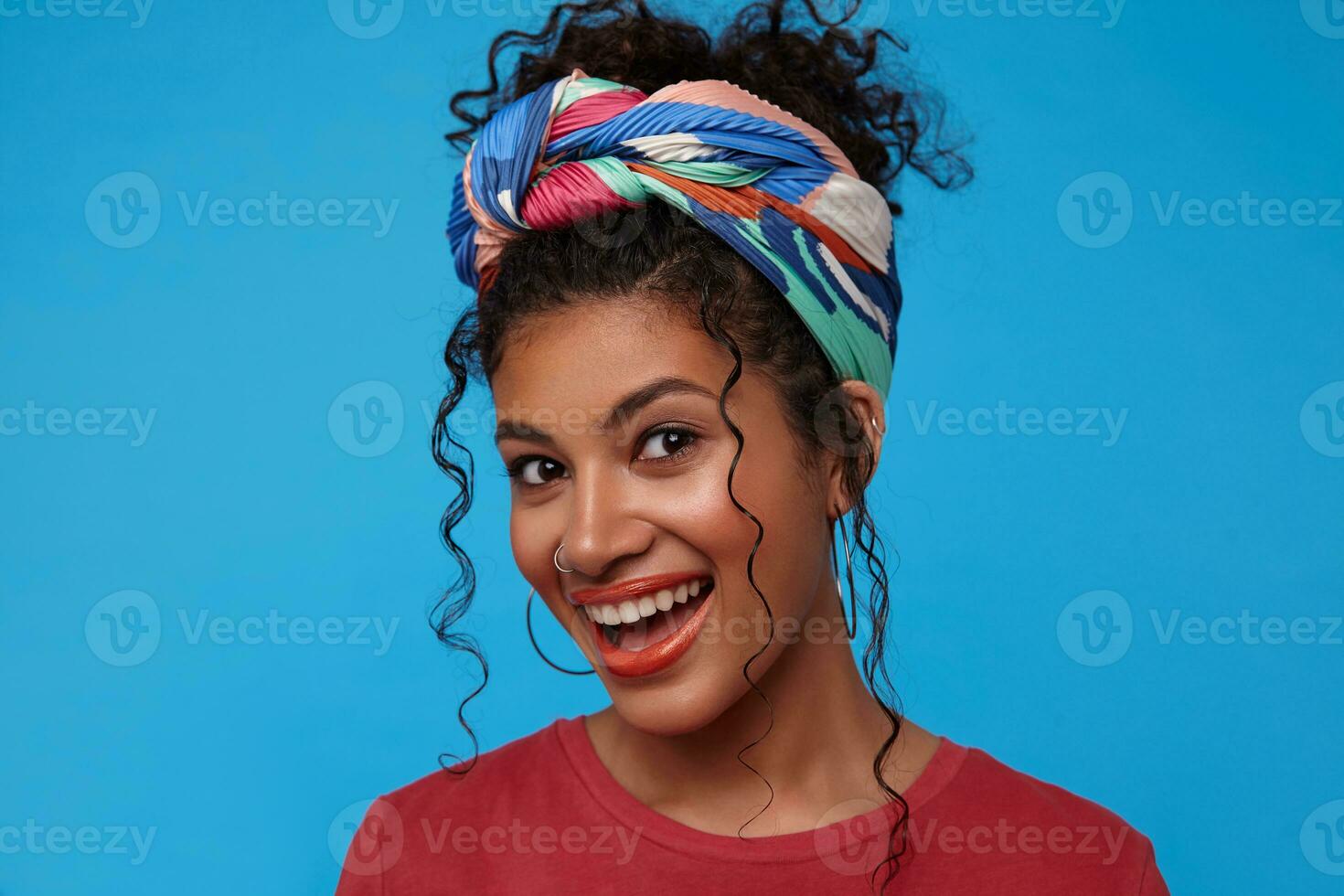 Portrait of young cute brunette curly woman with festive makeup being in nice mood and smiling happily while standing over blue background in colored clothes photo