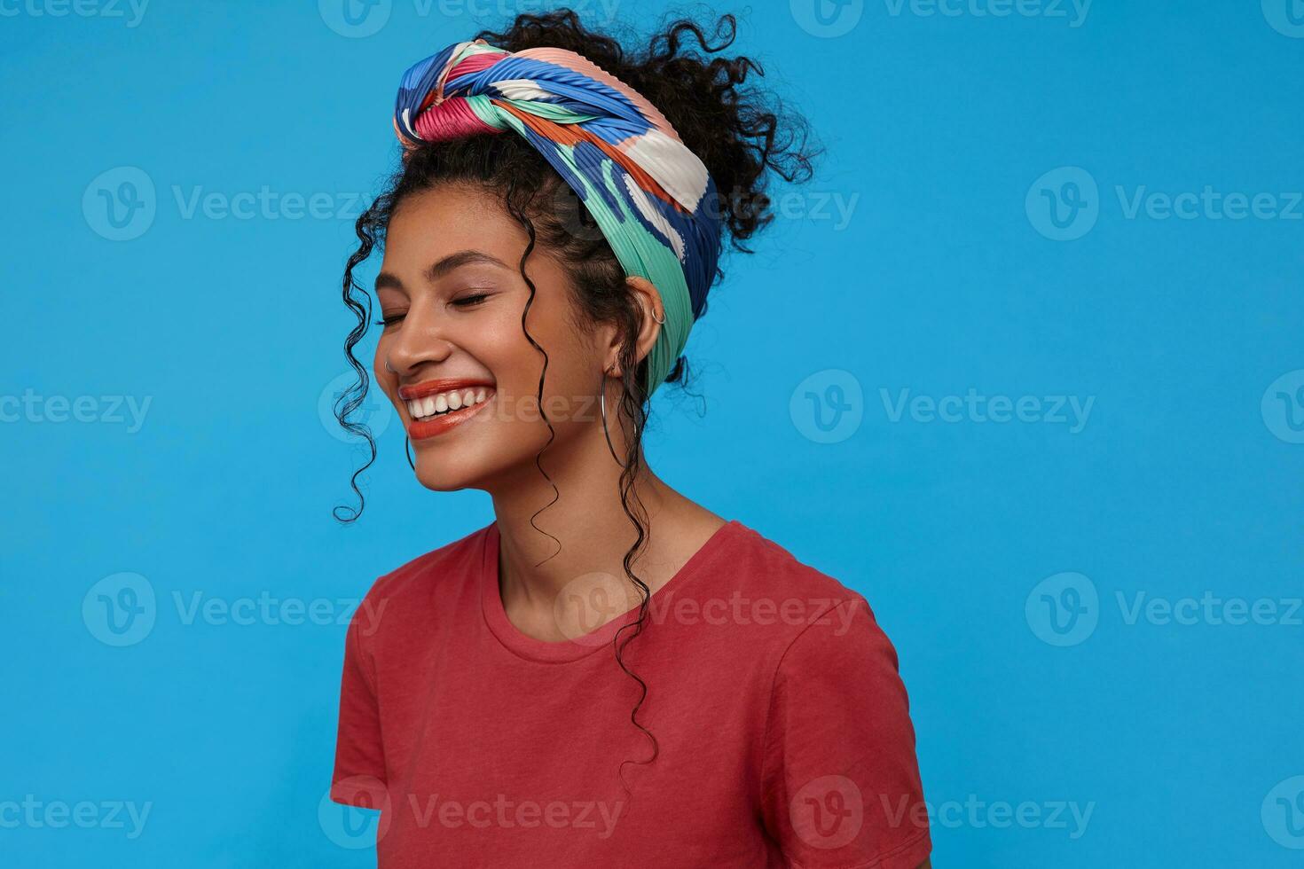 Good looking happy young brunette curly woman with multi-colored headband being in nice mood and miling gladly with closed eyes while standing over blue background photo