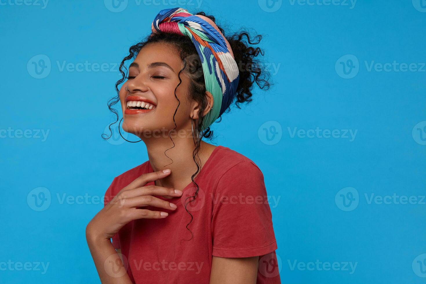 Indoor photo of young cheerful dark haired woman keeping raised hand on her chest and laughing happily with closed eyes, isolated over blue background