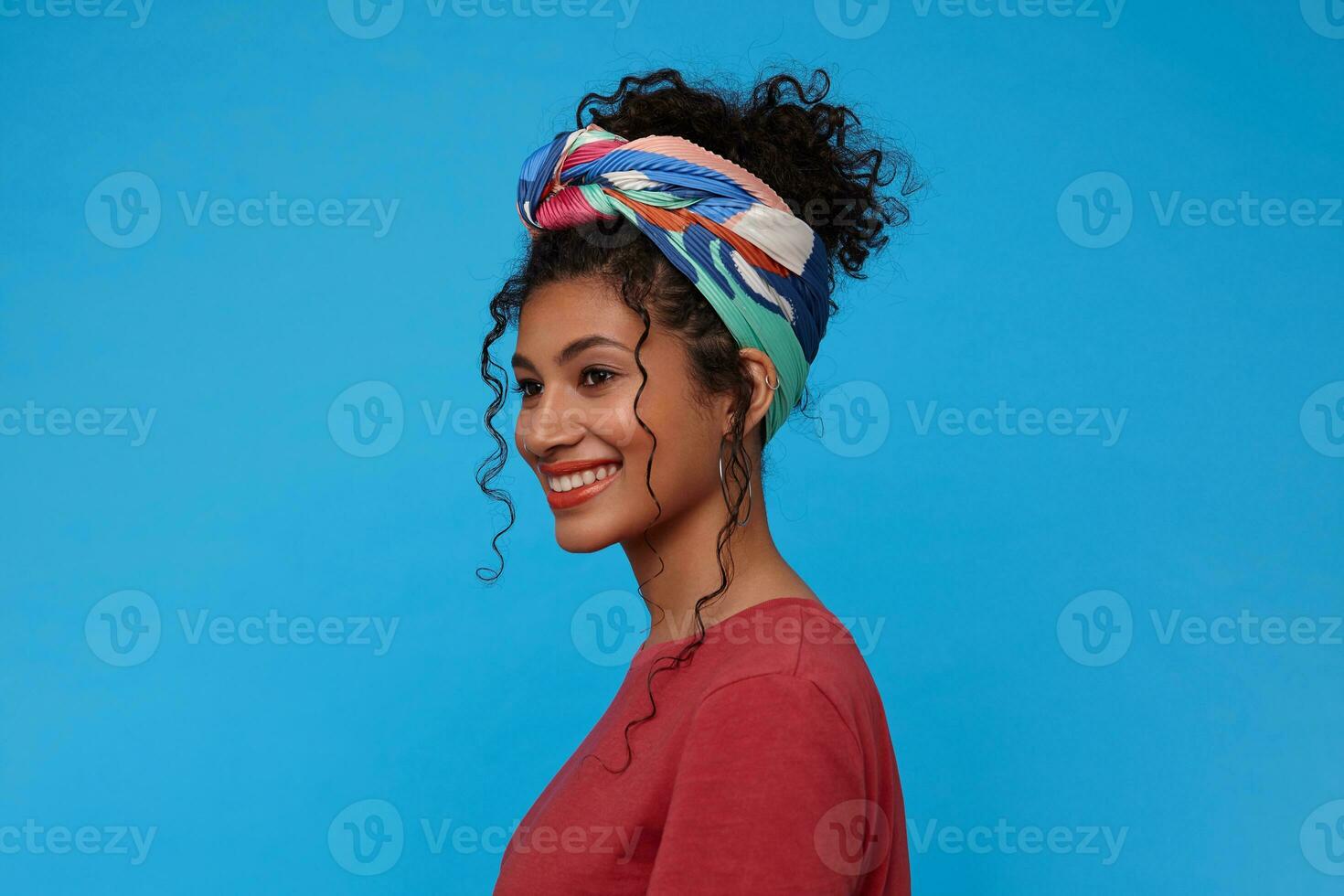 Side view of young cheerful brunette curly woman with festive makeup smiling broadly while looking gladly ahead, isolated over blue background in casual clothes photo