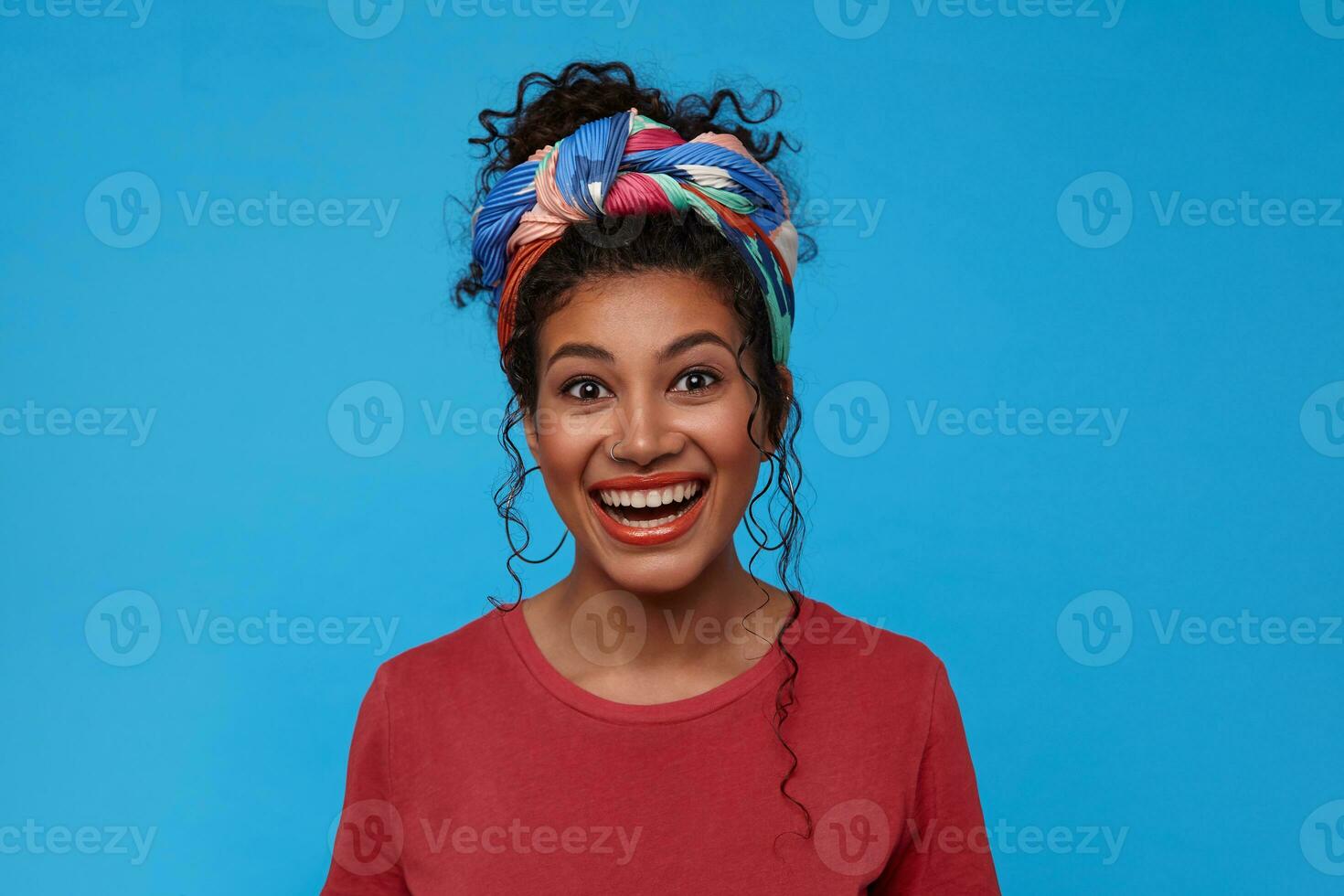 Indoor photo of young pretty dark haired curly woman with gathered hair smiling broadly while looking joyfully at camera, isolated over blue background
