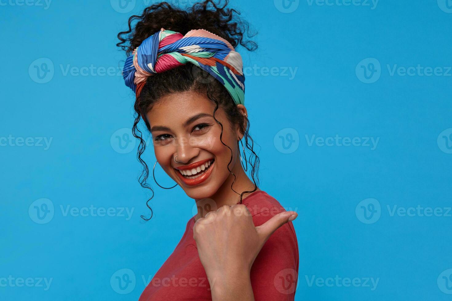 Portrait of young beautiful dark haired curly female with festive makeup thumbing back with raised hand and smiling happily at camera, standing over blue background photo