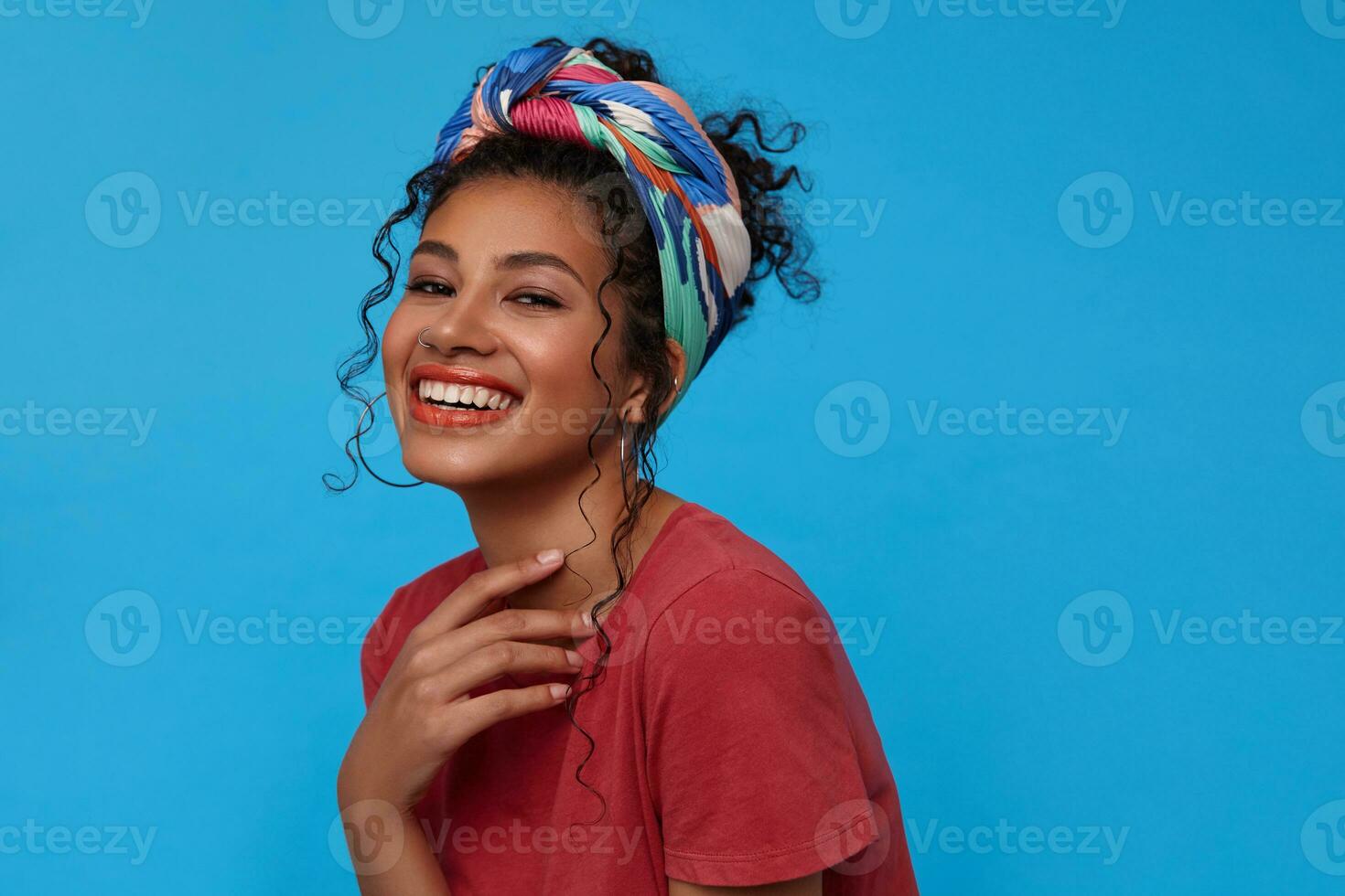 Studio photo of young attractive joyful brunette curly lady keeping her hand raised and looking gladly at camera with broad smile, isolated over blue background