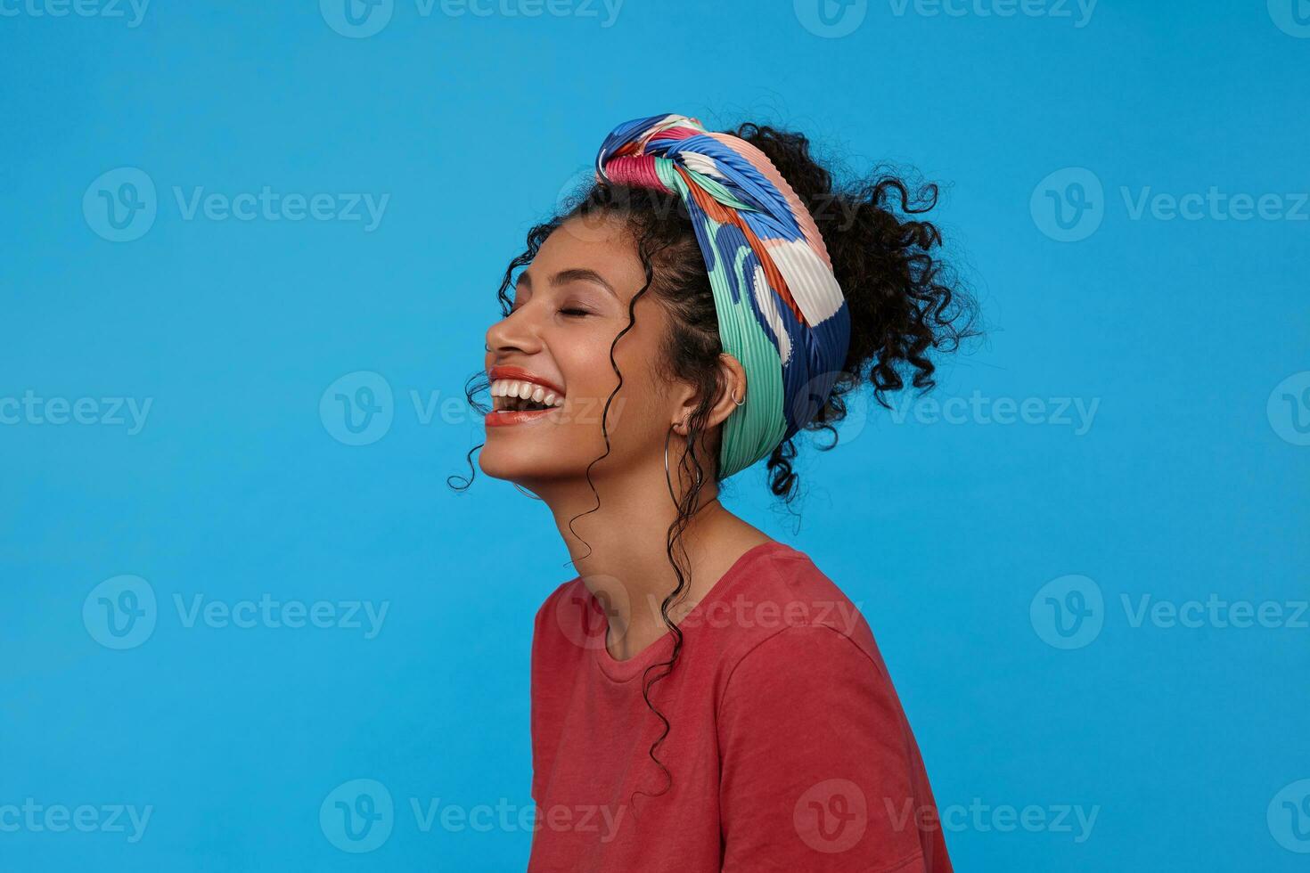 Pleasant looking young cheerful dark haired curly lady throwing back her head while laughing happily with closed eyes, standing over blue background with hands down photo