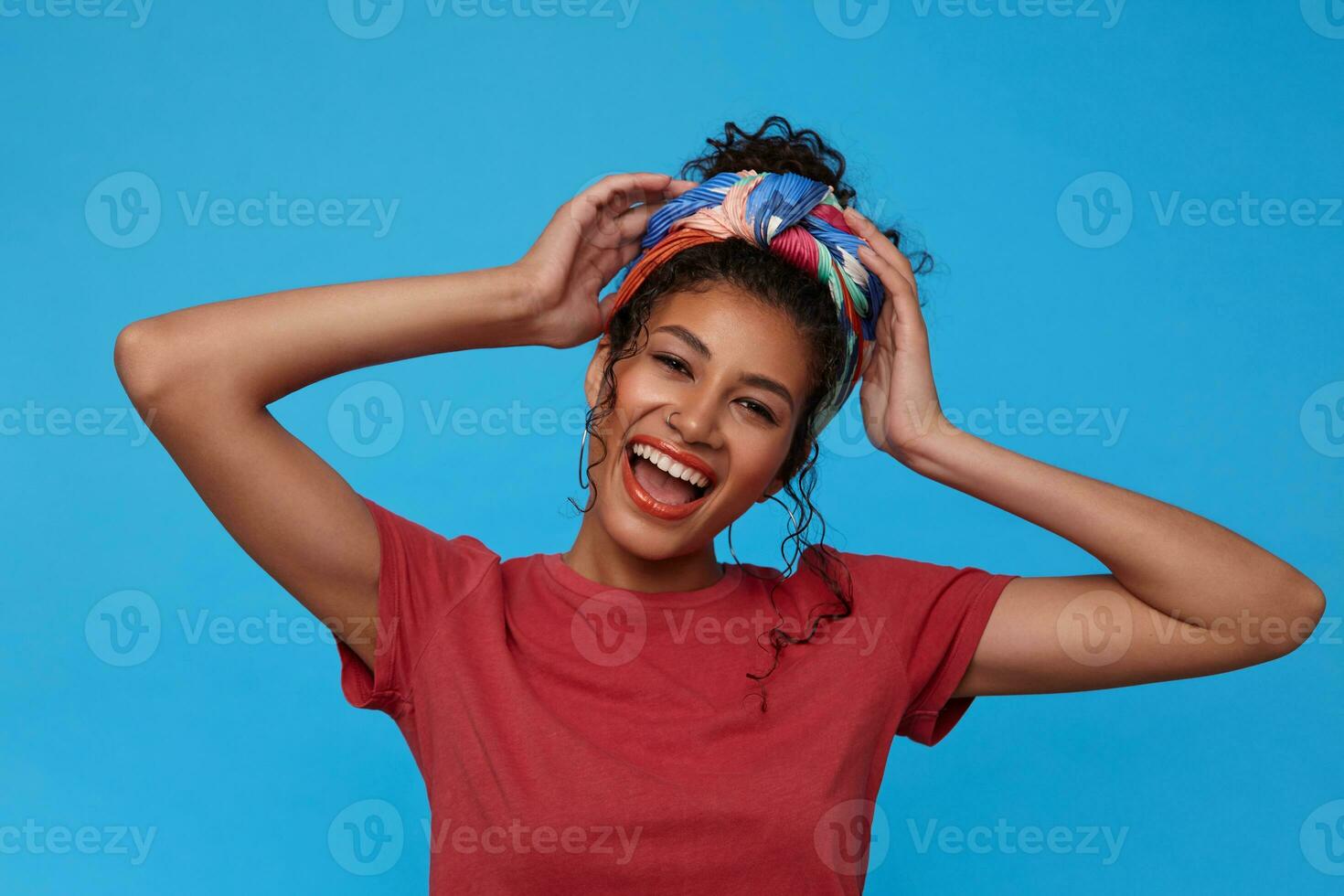 Portrait of young lovely dark haired curly female with festive makeup laughing happily and raising emotionally hands to her head, isolated over blue background photo