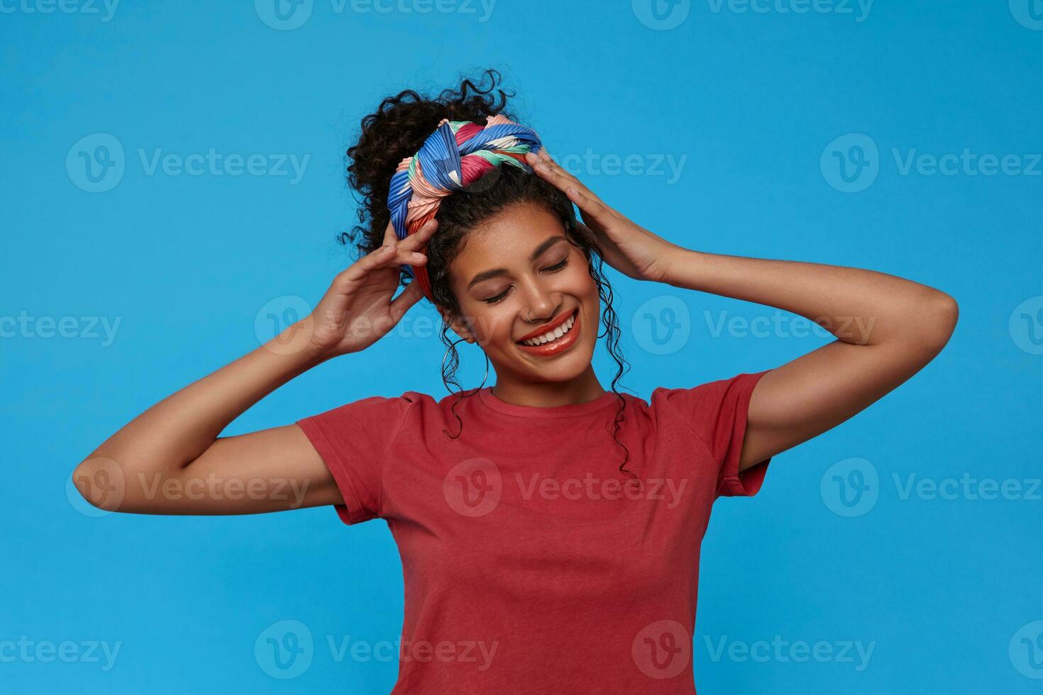 Pleased young cheerul brunette curly woman with gathered hair holding hands on her head and smiling widely with closed eyes, isolated over blue background photo