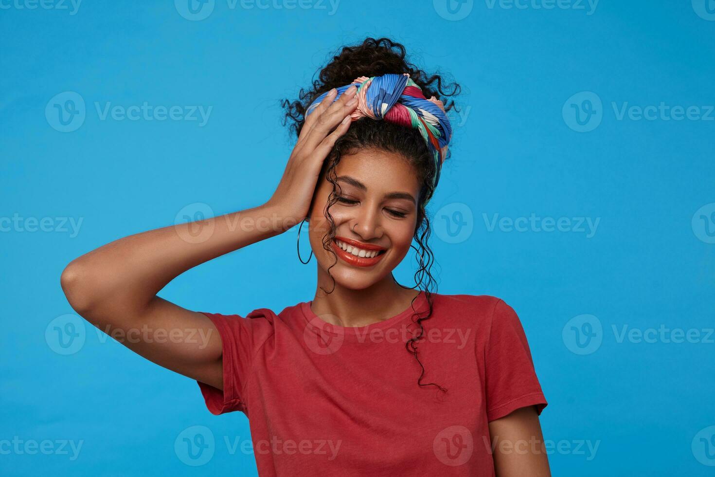 Pleasant looking young glad dark haired curly lady holding raised palm on her head and smiling happily with closed eyes, standing over blue background photo