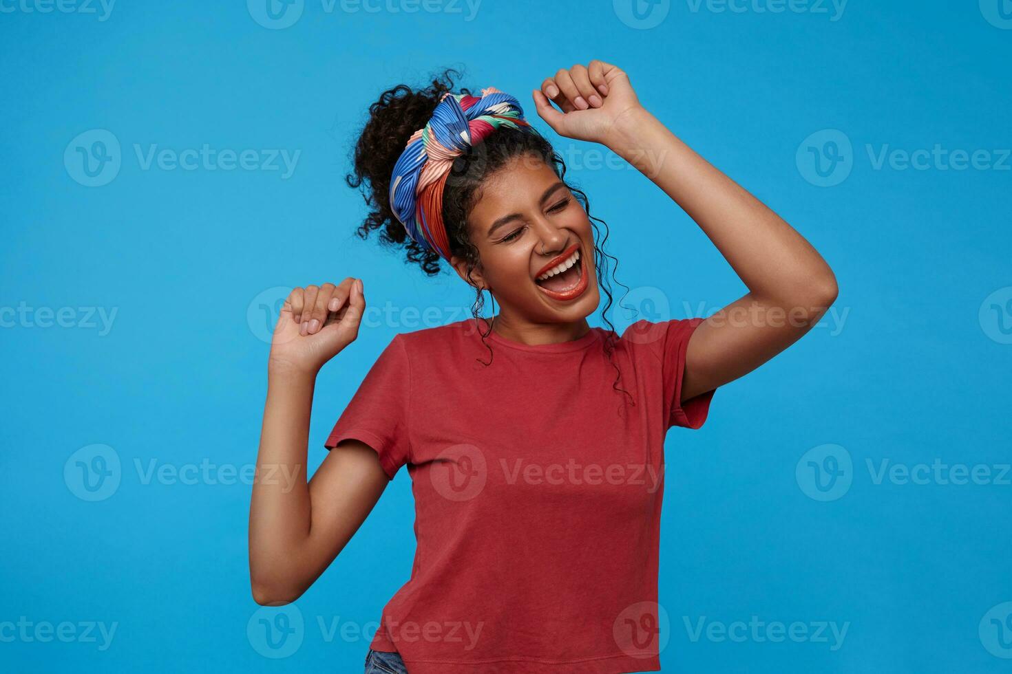 Joyful young attractive brunette curly woman with multi-colored headband singing and dancing cheerfully while standing over blue background with raised hands photo