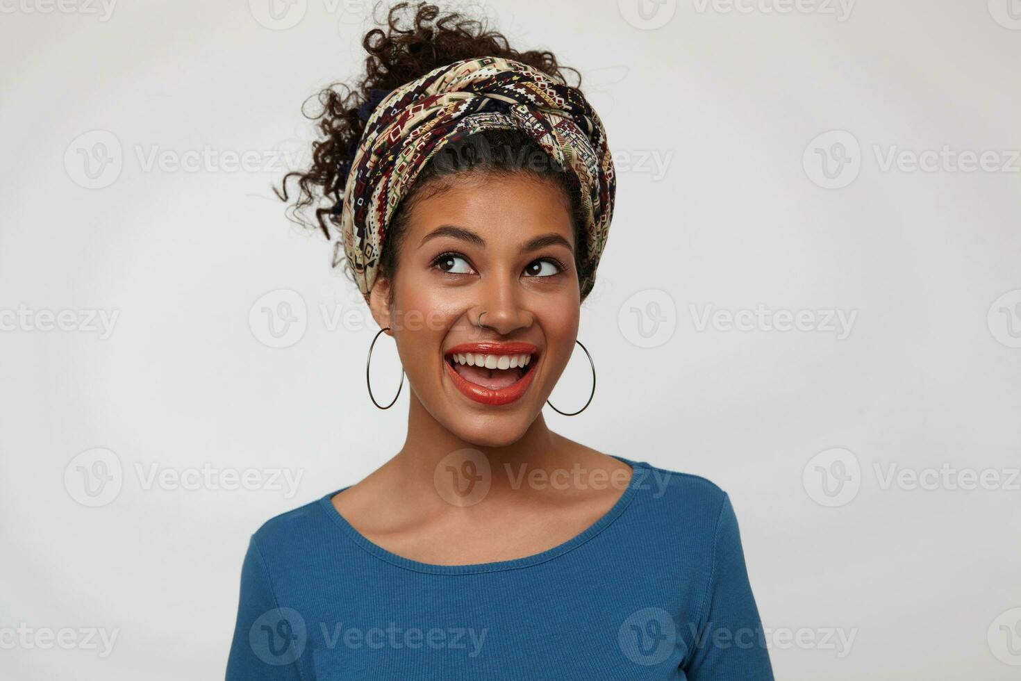 Portrait of agitated young brunette woman with gathered curly hair looking emotionally upwards with broad smile, standing over white background in colored wear photo