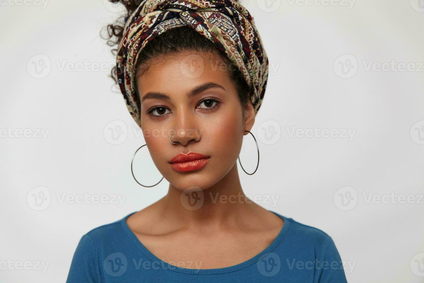 Portrait of young pretty brunette dark skinned female dressed in colored clothes keeping her lips folded while looking calmly at camera, isolated over white background photo