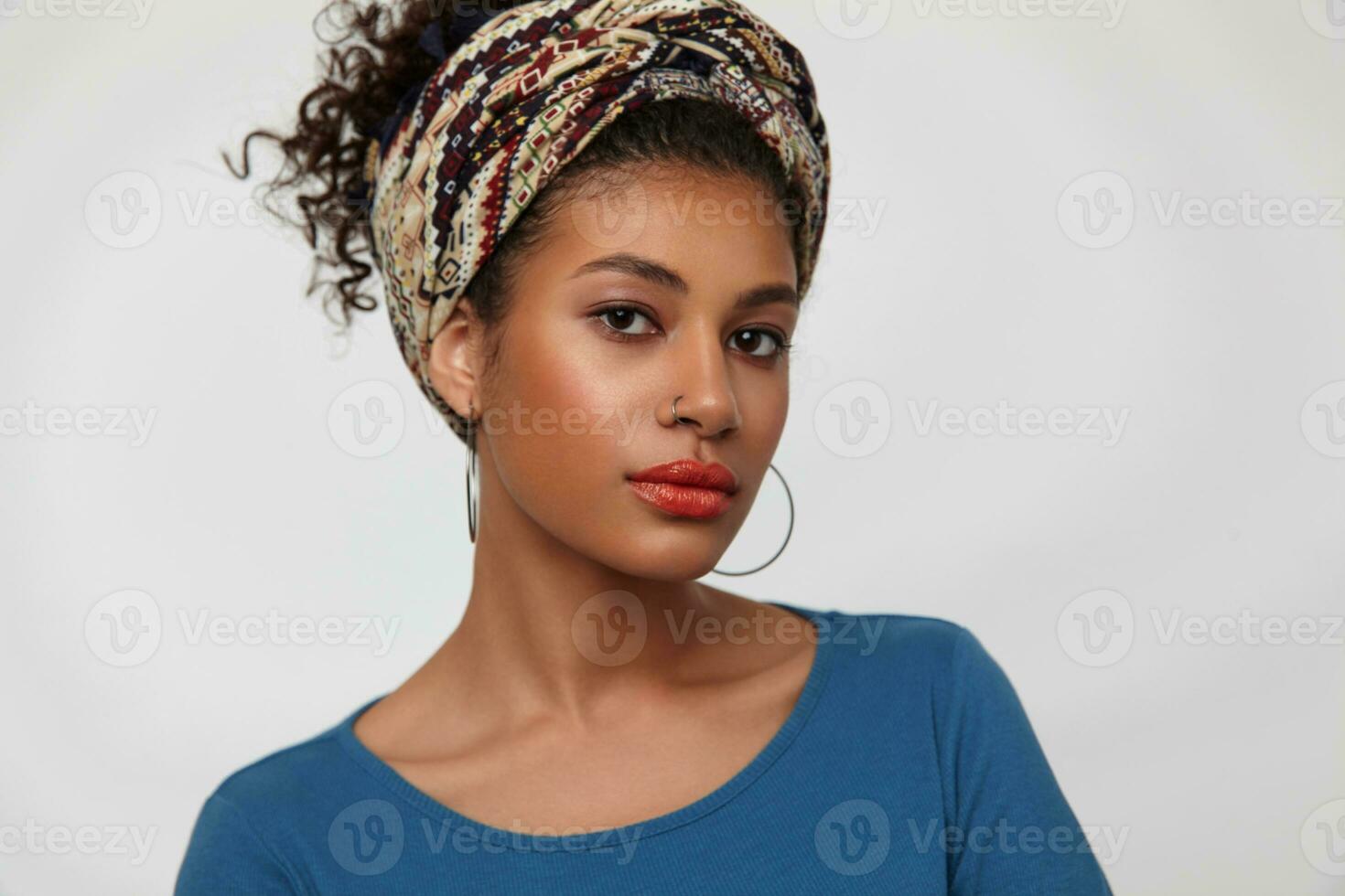 Portrait of pensive young attractive curly brunette woman with nose piercing looking attentively at camera and keeping lips folded, standing over white background photo