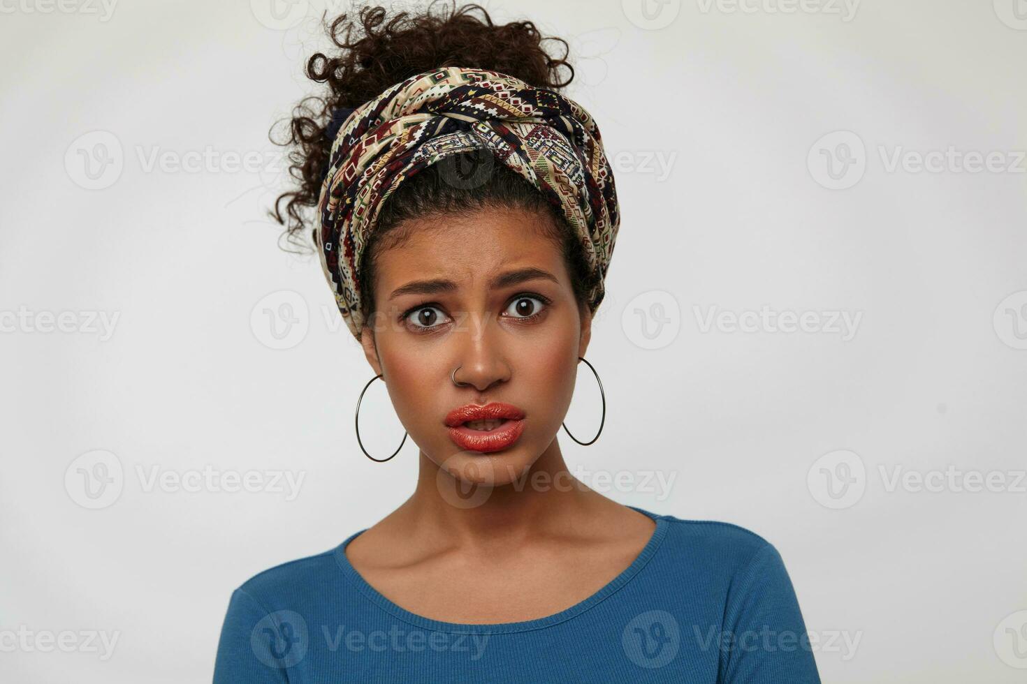 Bewildered young attractive brunette woman with casual hairstyle rounding her brown eyes while looking confusedly at camera, standing over white background photo