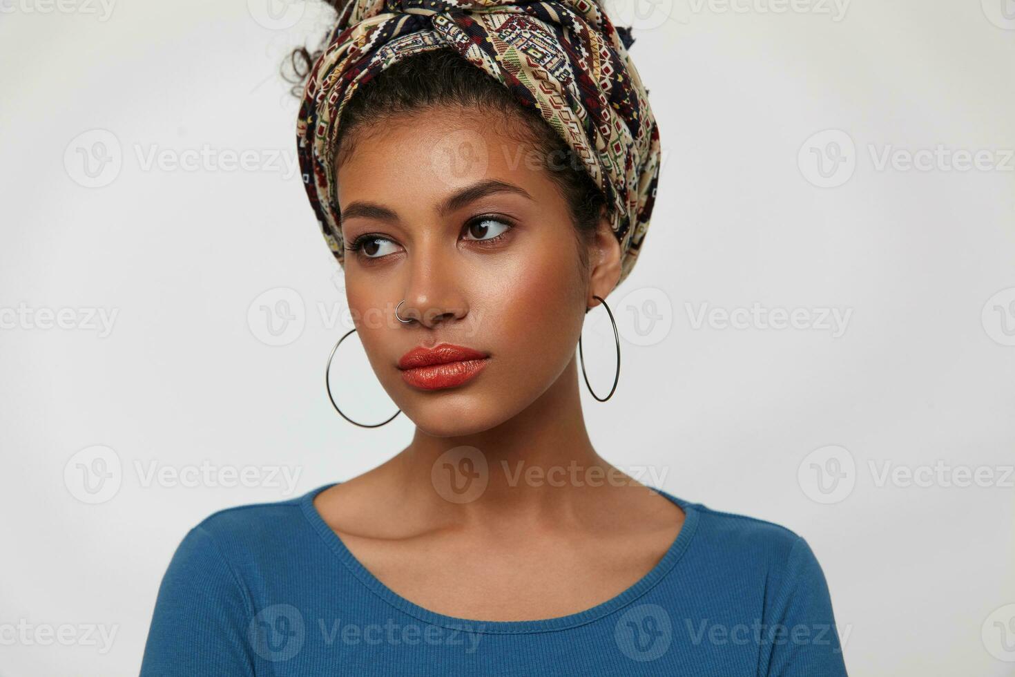 Close-up of young beautiful dark haired curly woman with gathered curly hair looking attentively aside while standing over white background with hands down photo