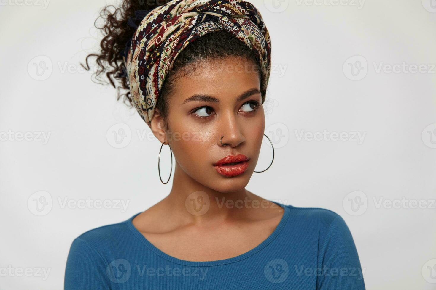 Bewildered young pretty dark haired curly lady with casual hairstyle looking wonderingly aside while standing over white background in colored clothes photo