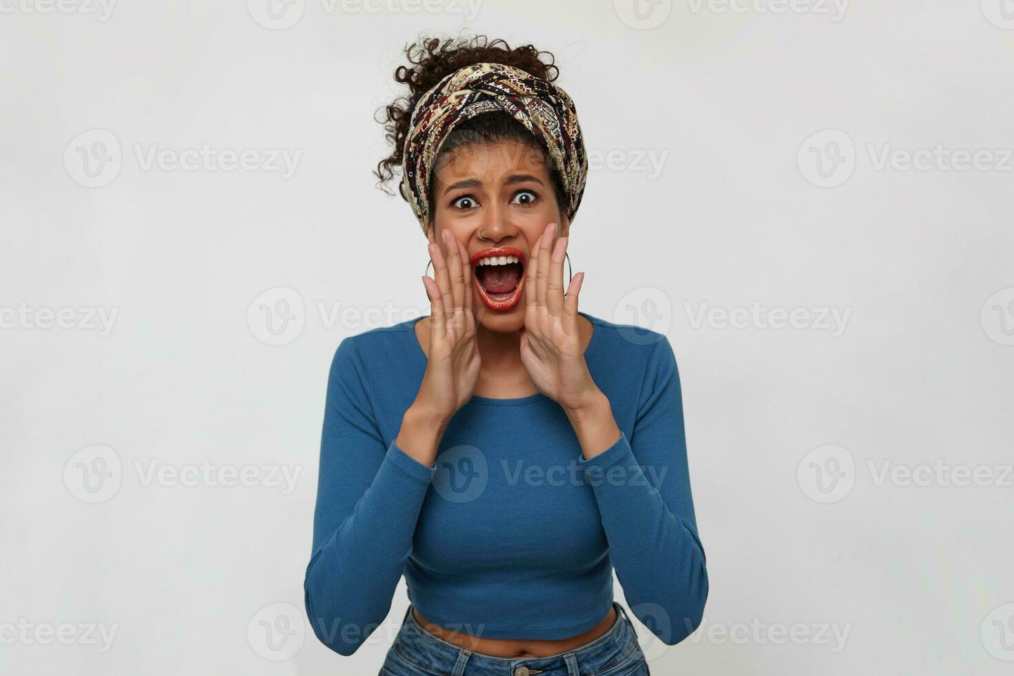 Agitated young dark haired curly woman with dark skin raising hands to her mouth while screaming excitedly with wide mouth opened, isolated over white background photo