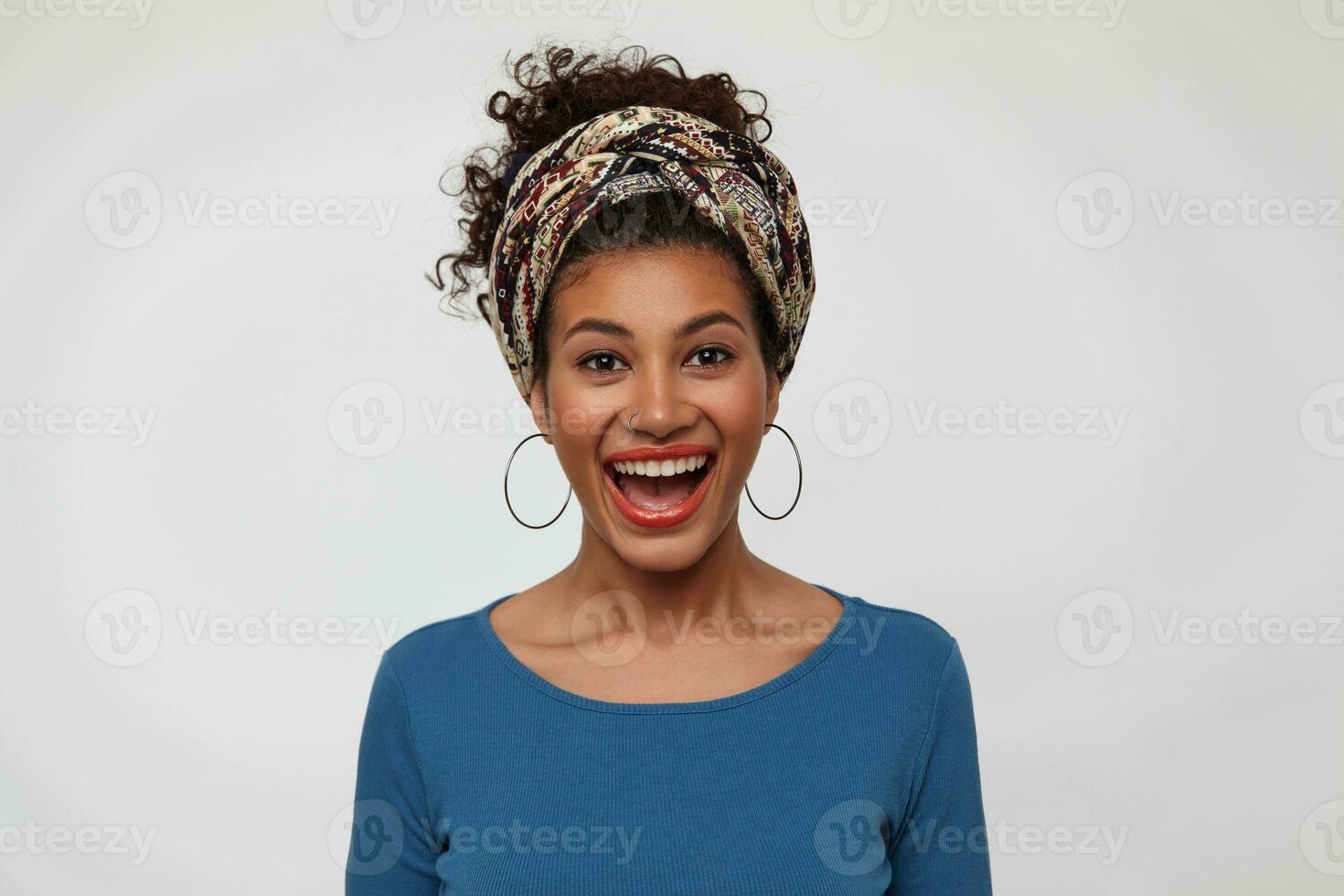 Studio photo of happy young dark haired curly trendy lady laughing cheerfully while rejoicing about something, isolated over white background with hands down