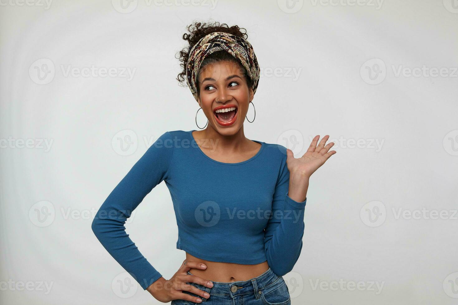 Studio shot of young joyful dark haired curly lady dressed in trendy colored clothes raising hand while looking cheerfully aside with wide mouth opened, isolated over white background photo