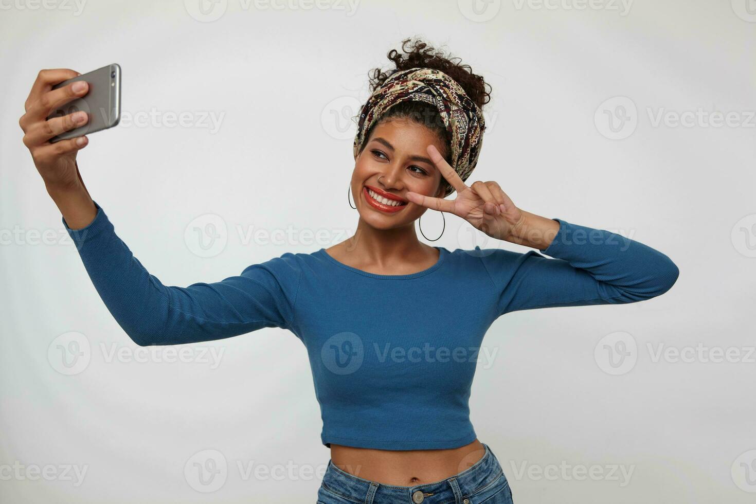Cheerful young lovely dark haired curly lady with nose piercing smiling gladly and raising victory gesture to her face while making selfie, isolated over white background photo