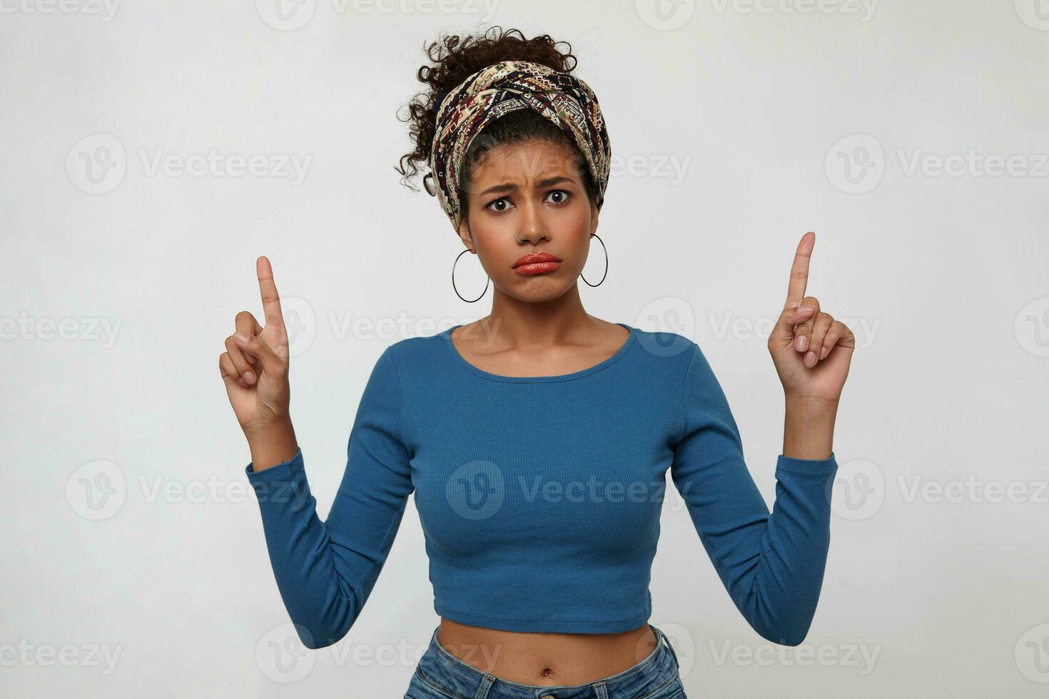 Bewildered young pretty brunette dark skinned lady folding her lips and frowning eyebrows while pointing confusedly upwards, standing over white background photo