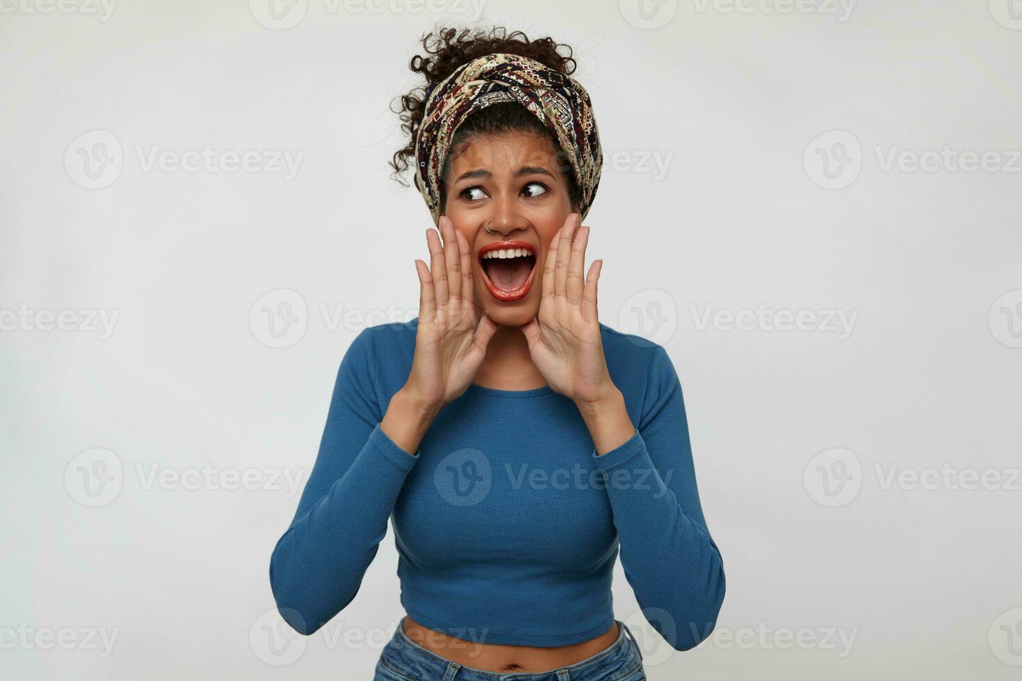 Excited young pretty brunette dark skinned woman with gathered hair keeping raised hands near her mouth while shouting with opened mouth, isolated over white background photo