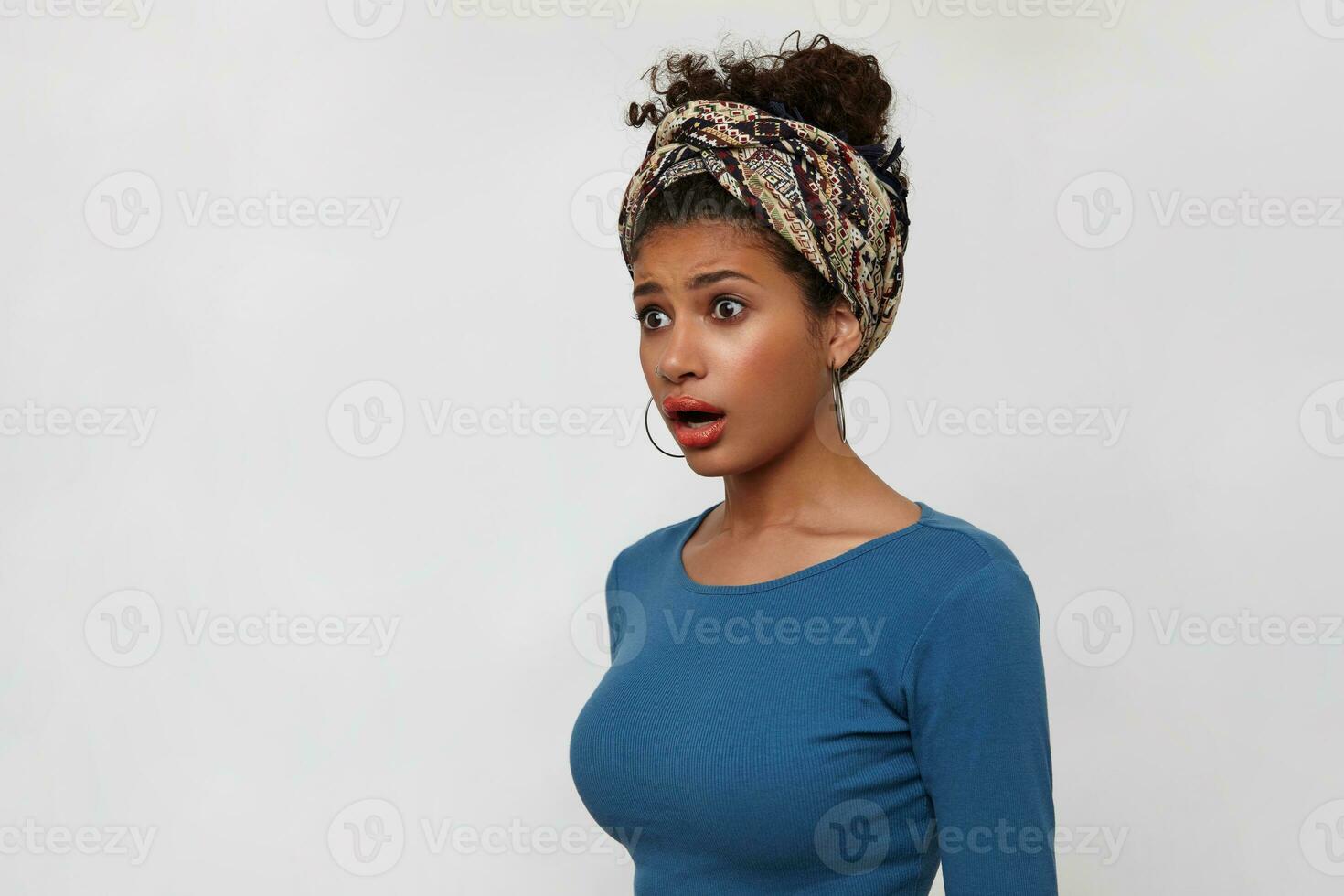 Bemused young dark haired curly female with gathered hair looking dazedly aside with wide eyes and mouth opened, standing over white background with hands down photo