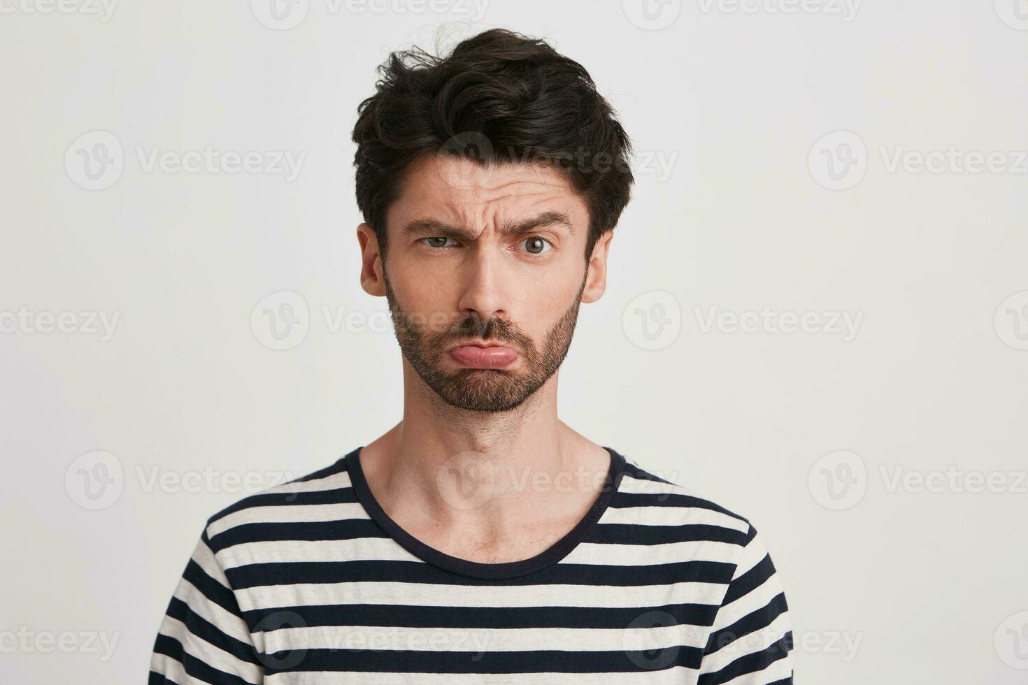 Closeup of sad offended young man with bristle wears striped t shirt feels upset and looks directly in camera isolated over white background photo