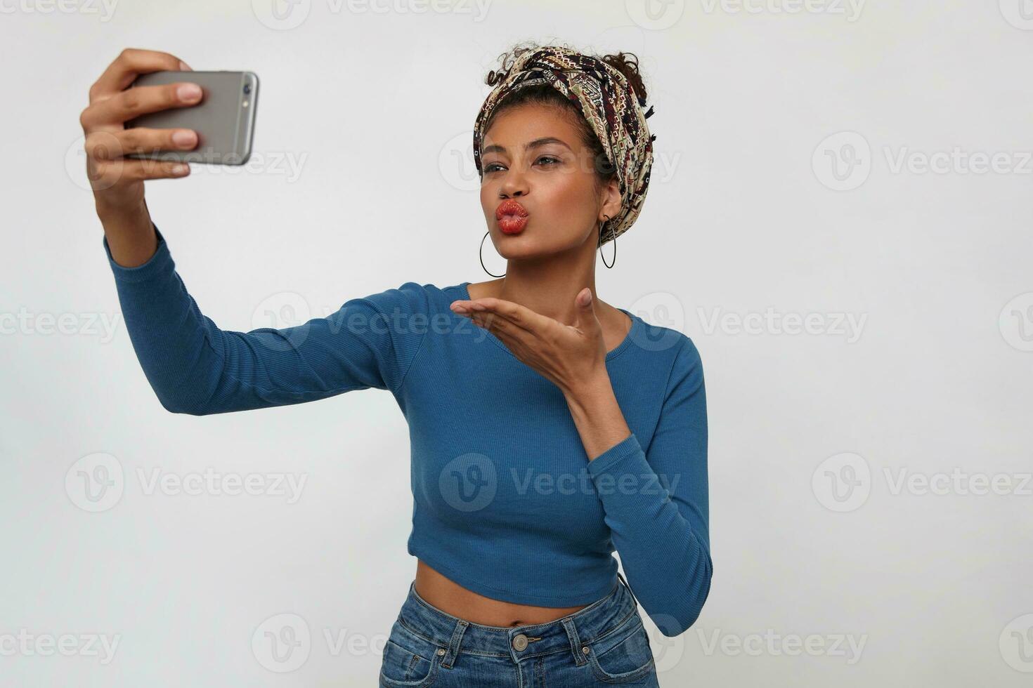 Attractive young brunette dark skinned female sending air kiss at camera while making photo of herself on mobile phone, standing over white background