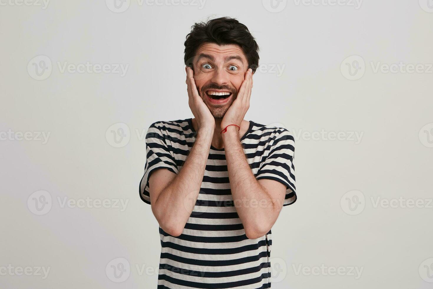 Indoor shot of optimistic male with glad expression, keeps hands on cheeks, feels shoked and happy, isolated over white wall photo
