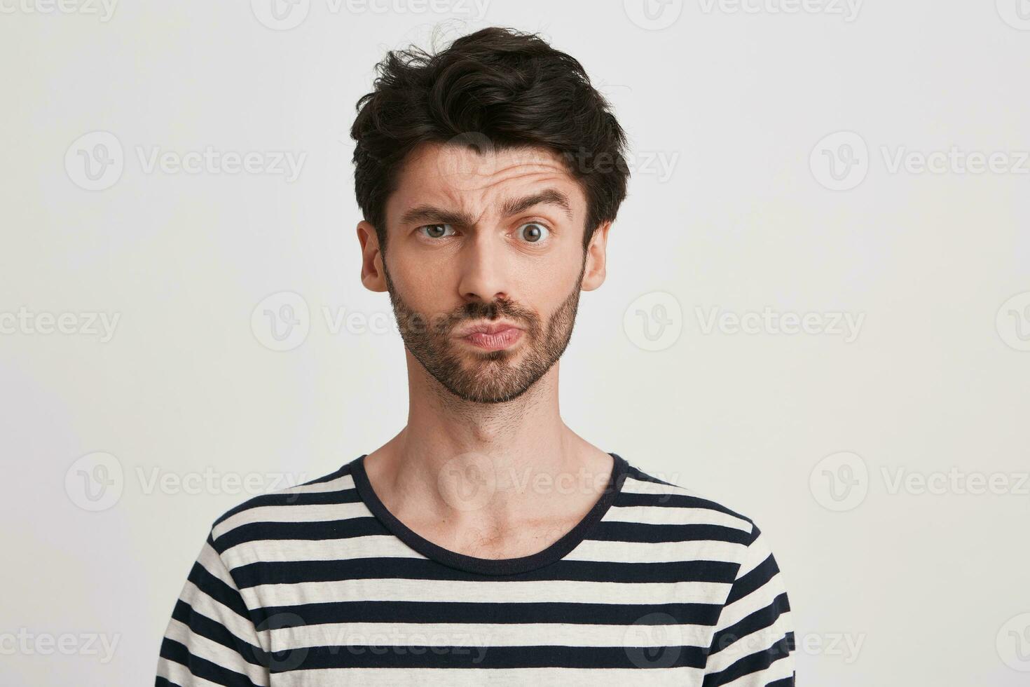 Portrait of pensive confused young man with bristle wears striped t shirt feels embarrassed and thinking isolated over white background Looks directly in camera photo