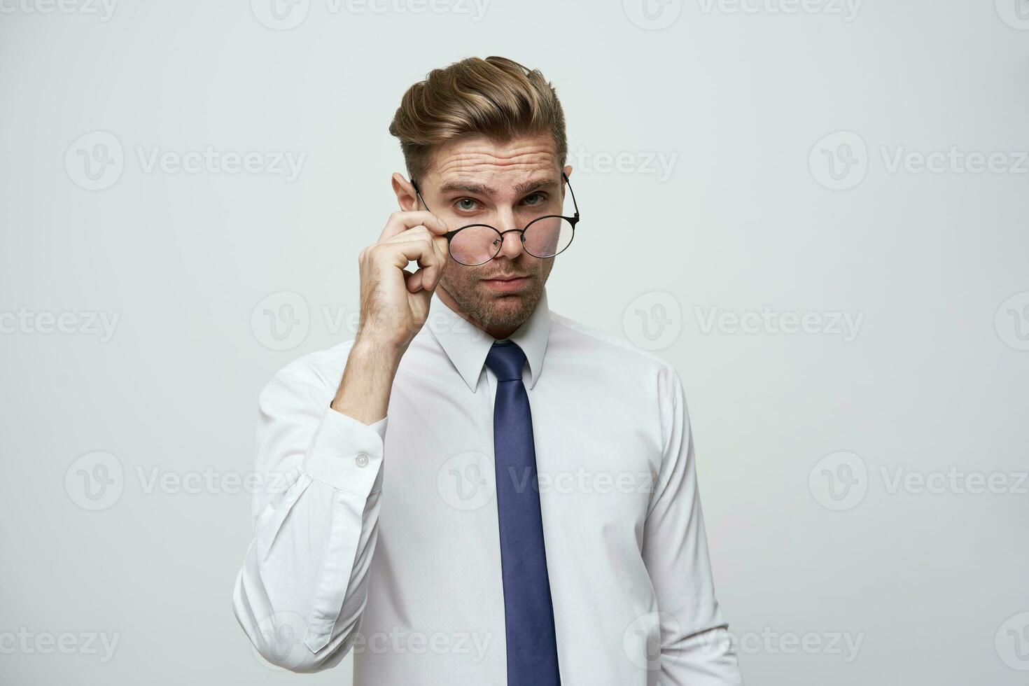 Attractive handsome businessman with stylish haircut, puts glasses down to the tip of the nose to see some details, dressed in perfectly ironed white shirt and blue tie,over white background photo