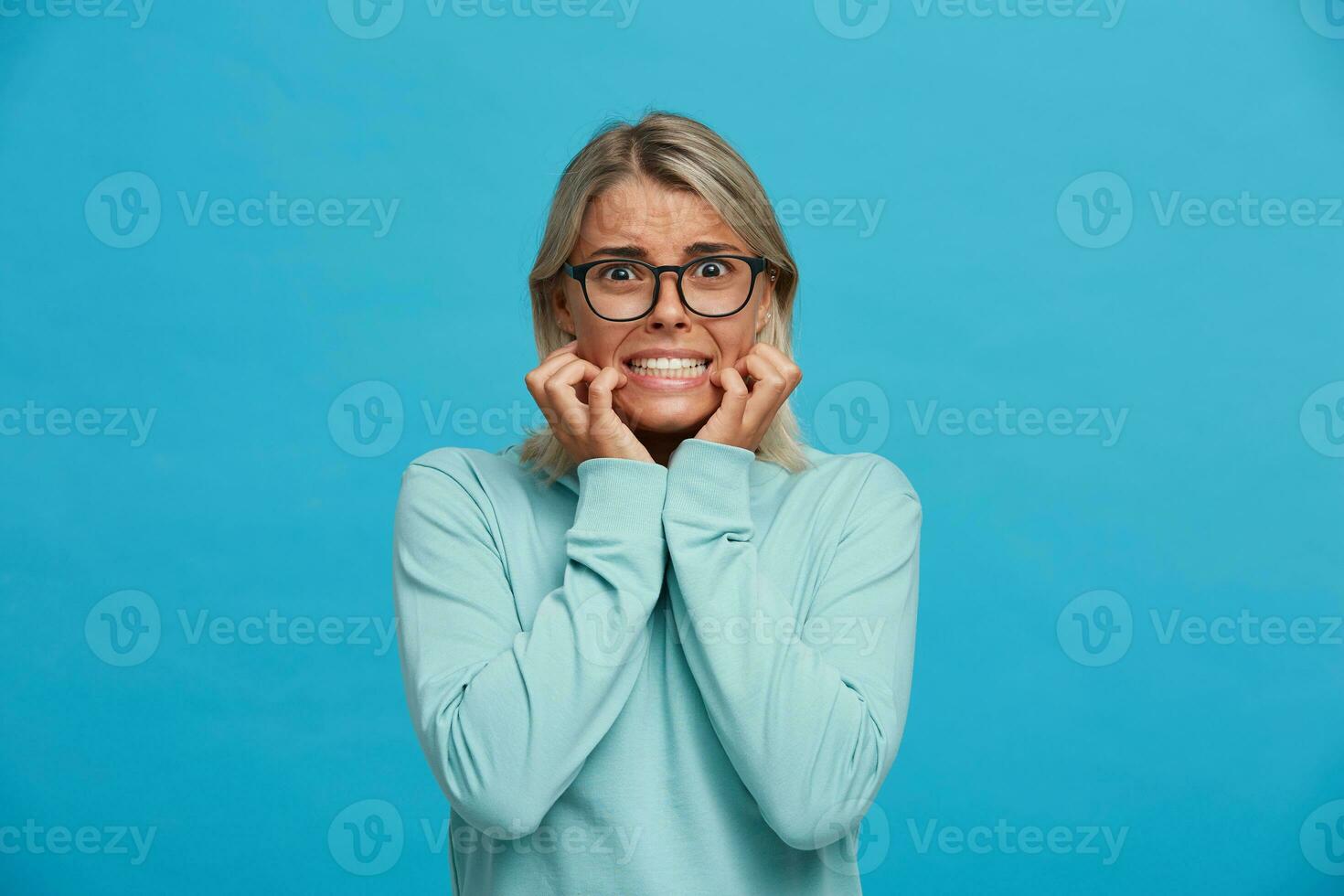 Portrait of scared blonde girl, looks frightened afraid chattering teeth with fear,bites nails from fright, hands holding cheeks, over blue background photo