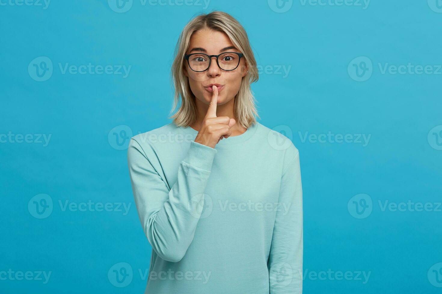 Emotional young girl in glasses calls for silence, keep a secret, do not tell anyone, keep quiet, do not make noise, keeps fore finger on lips, demonstrates silence gesture, on a blue background photo