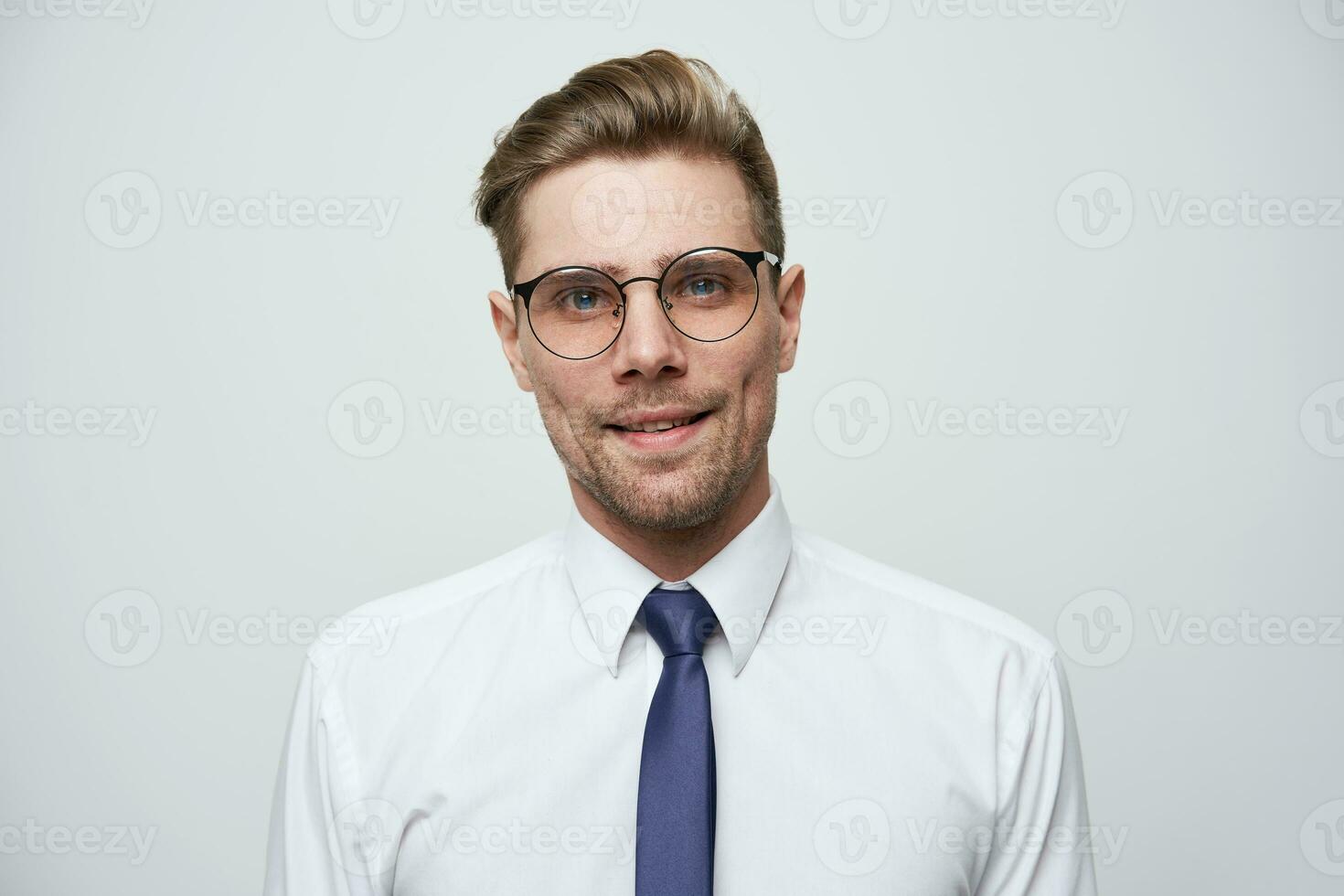 Young man with stylish haircut, in glasses,blue eyes look straight little smiling, feels calm and pleased, dressed in white shirt and blue tie, unshaven, over white background photo