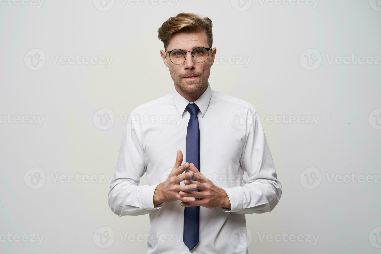 A young man stands with his fingers folded in front, looks through glasses, a little nerdy, tense, tries to make a choice or find a problem decision, on a white background photo