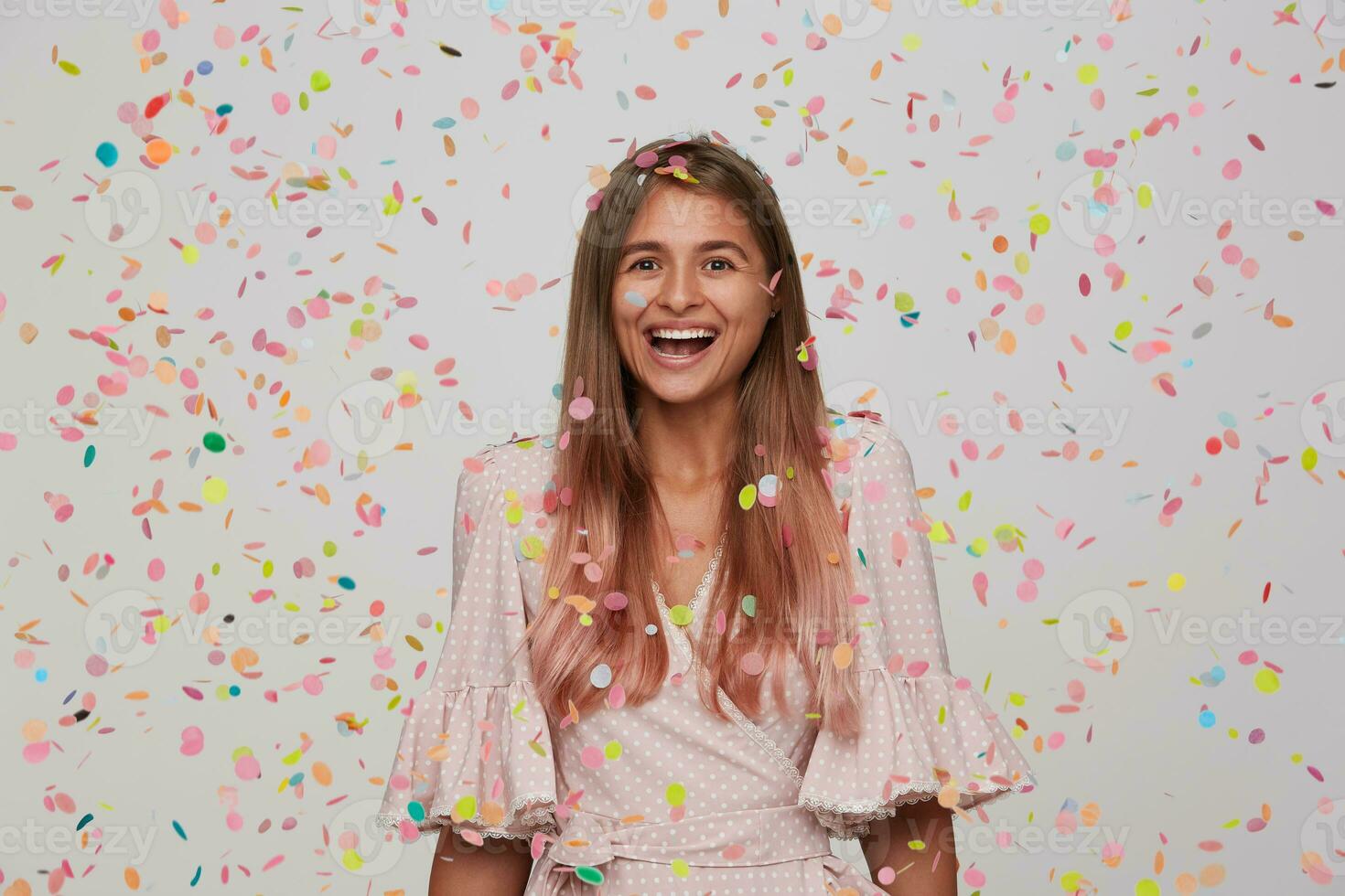 Portrait of happy lovely young woman with long dyed pastel pink hair wears polka dot pink dress laughing, celebrating and having party isolated over white background with confetti photo