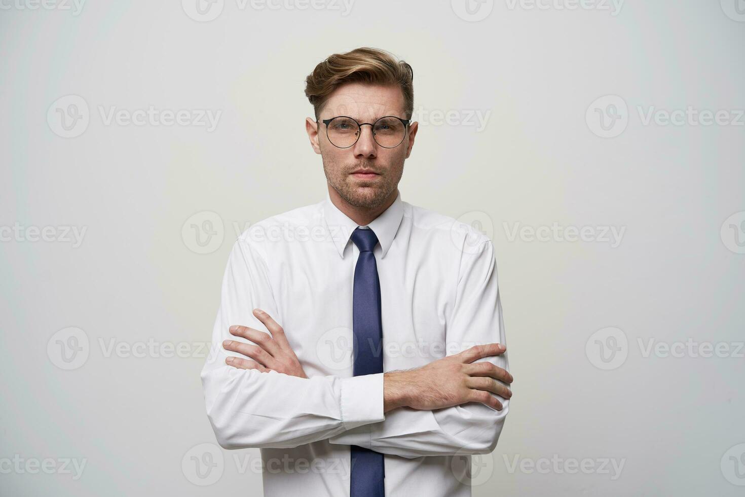 retrato de joven atractivo hombre mira evaluativo, refleja, ojos ligeramente estrechado, mira mediante anteojos, manos son cruzado, sin afeitar, vestido en blanco camisa y azul corbata, encima blanco antecedentes foto