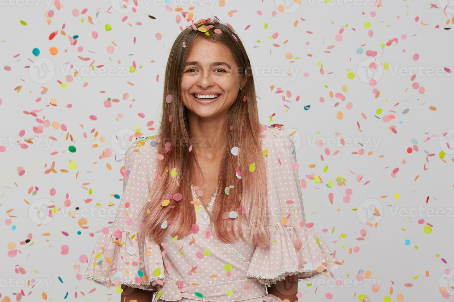 Surprised joyful charming young woman with long dyed pastel pink hair wears polka dot pink dress celebrating birthday, feels happy and laughing isolated over white background with confetti photo